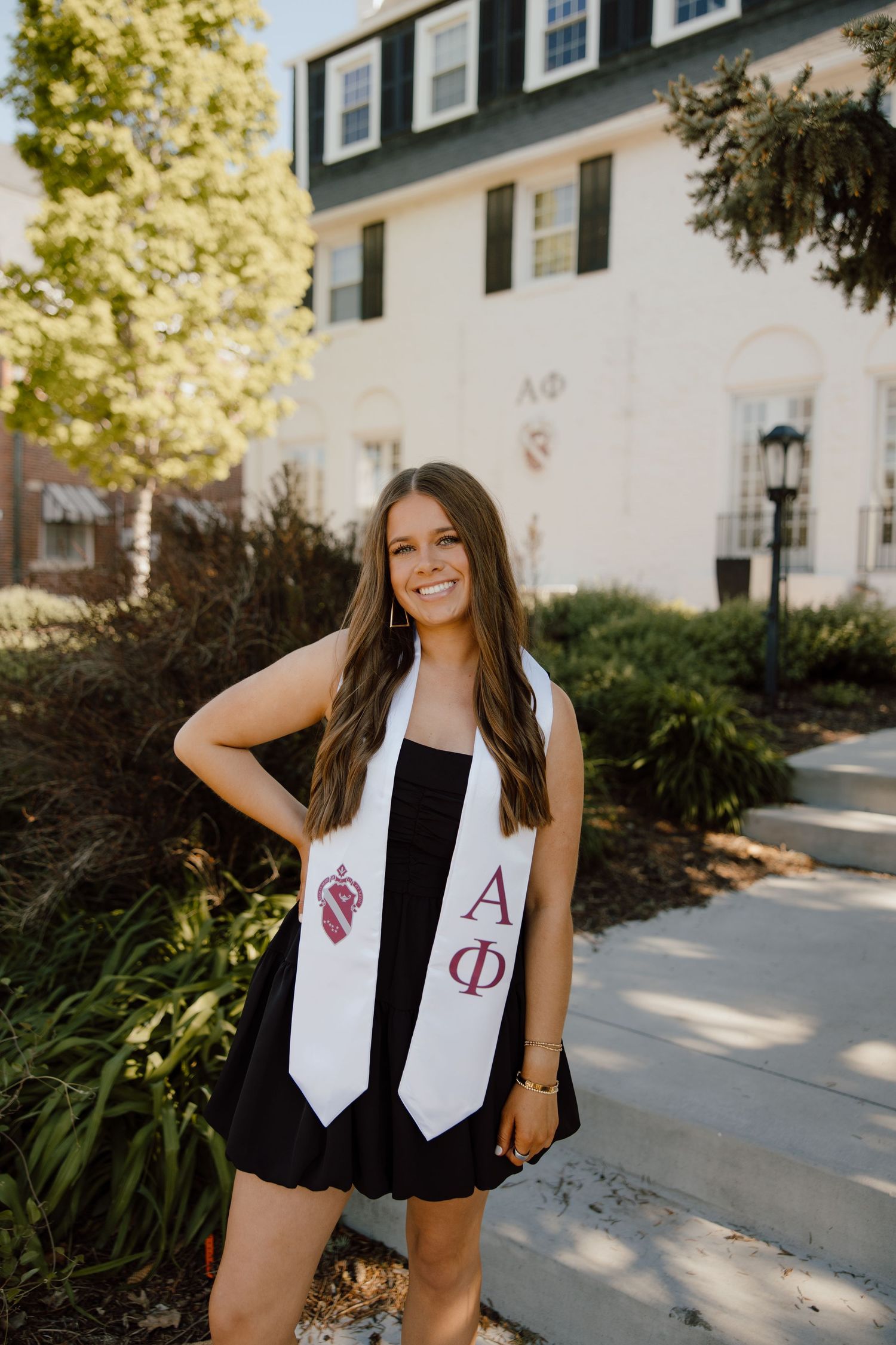 Graduate wearing black dress and white sorority stoles posing outside building.
