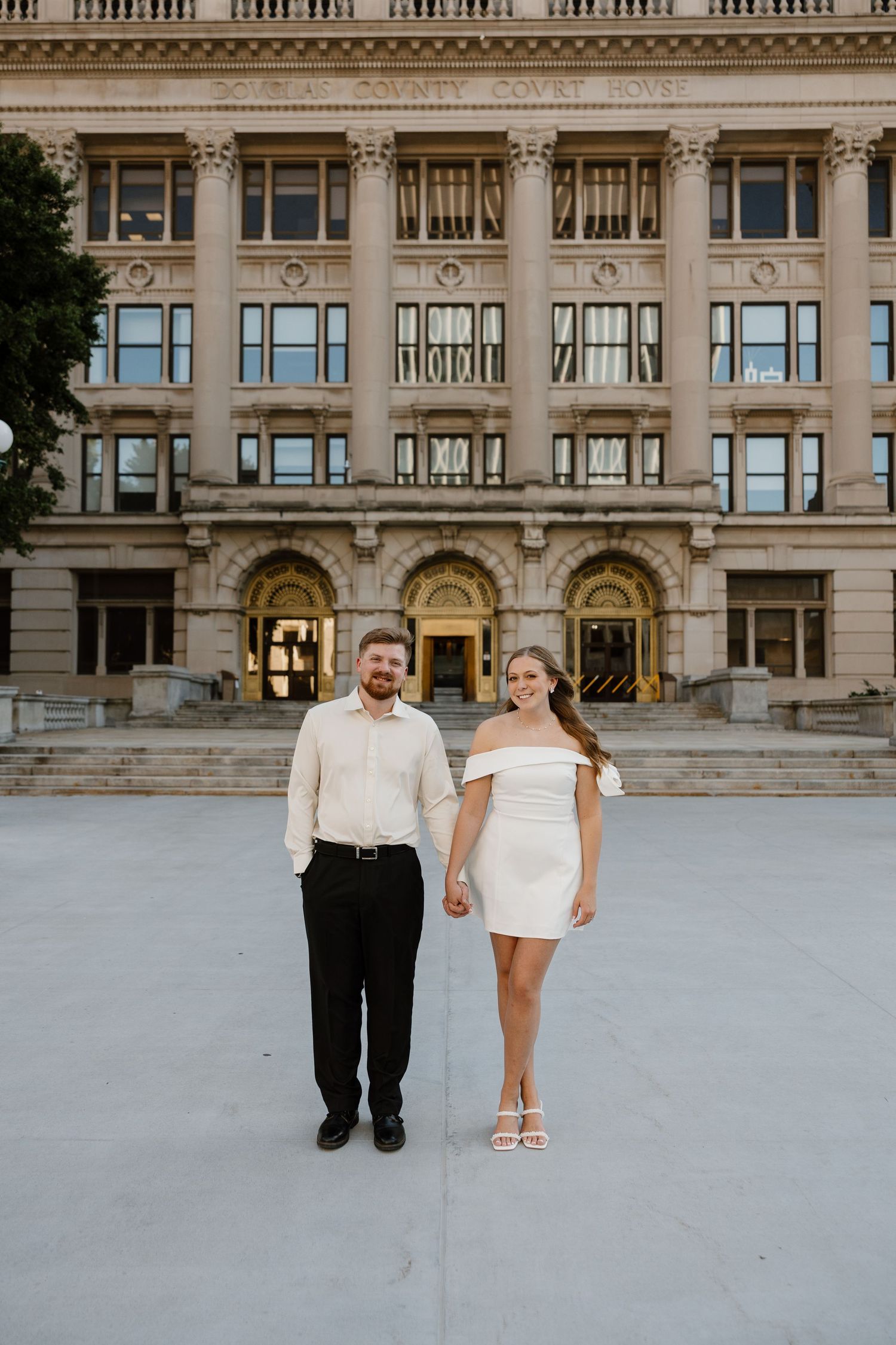 Couple poses together on wide plaza in front of ornate classical architecture.