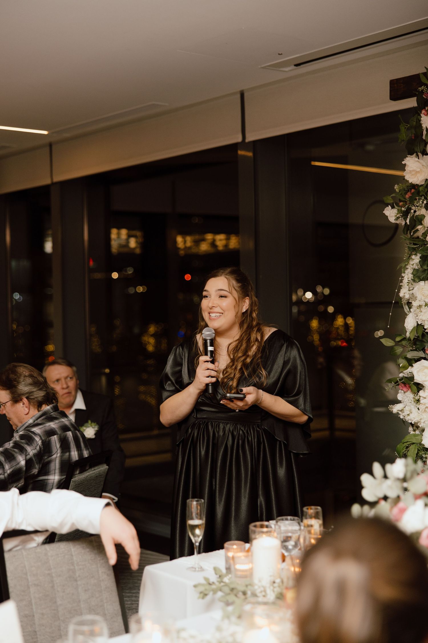 A wedding guest in black giving a speech at an elegant reception decorated with white floral arrangements.
