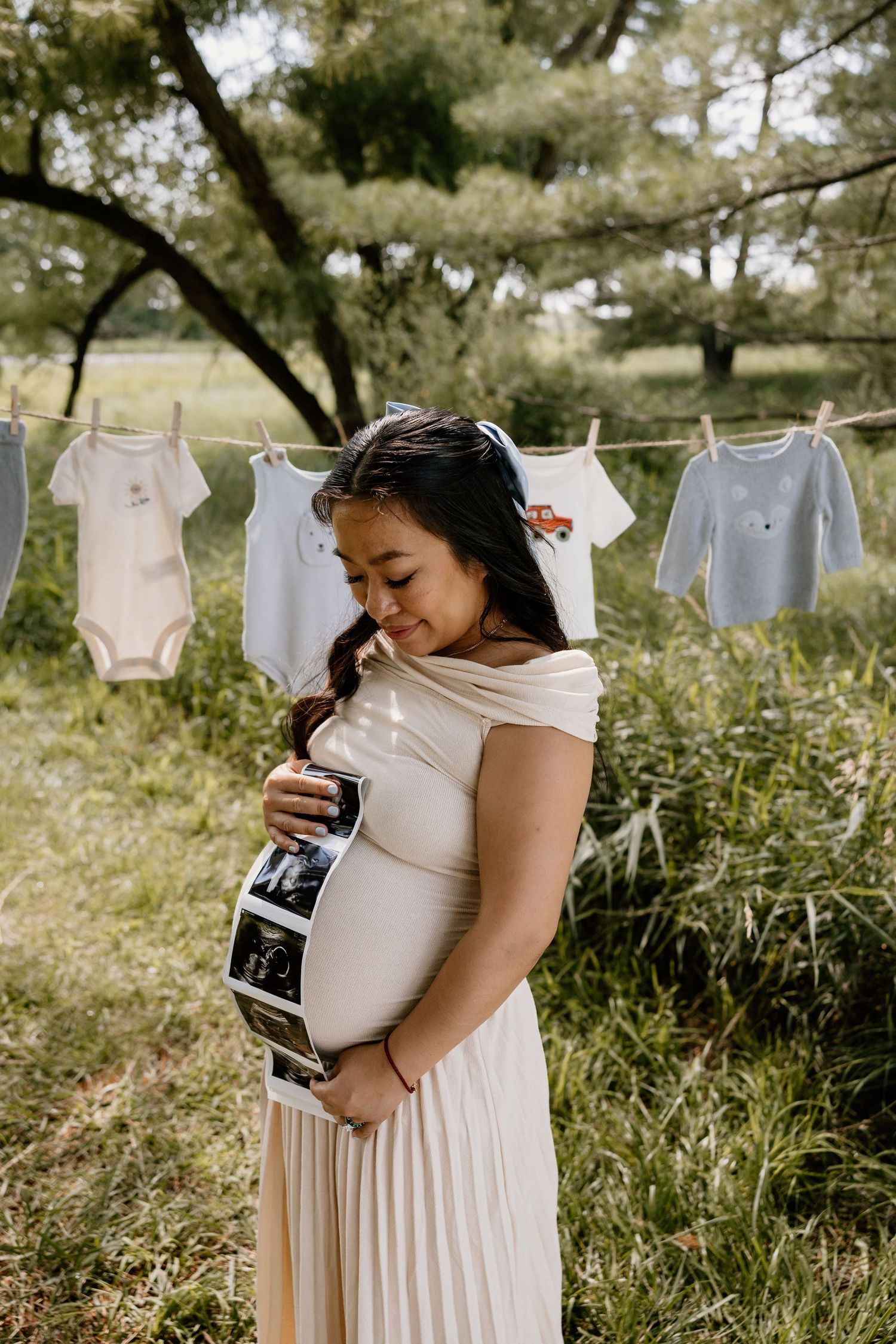 Maternity photoshoot featuring baby clothes on clothesline and ultrasound photos in natural setting.