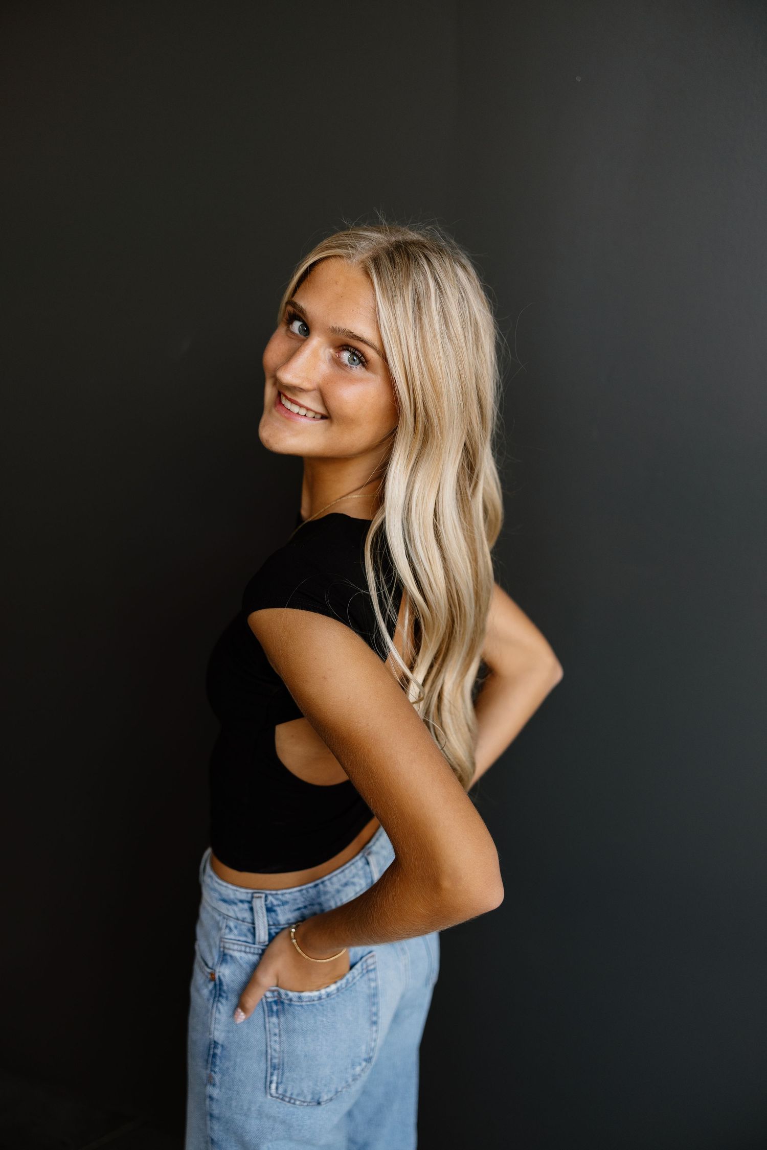 Woman with blonde hair poses against dark background in denim jeans and black top in artistic portrait series.