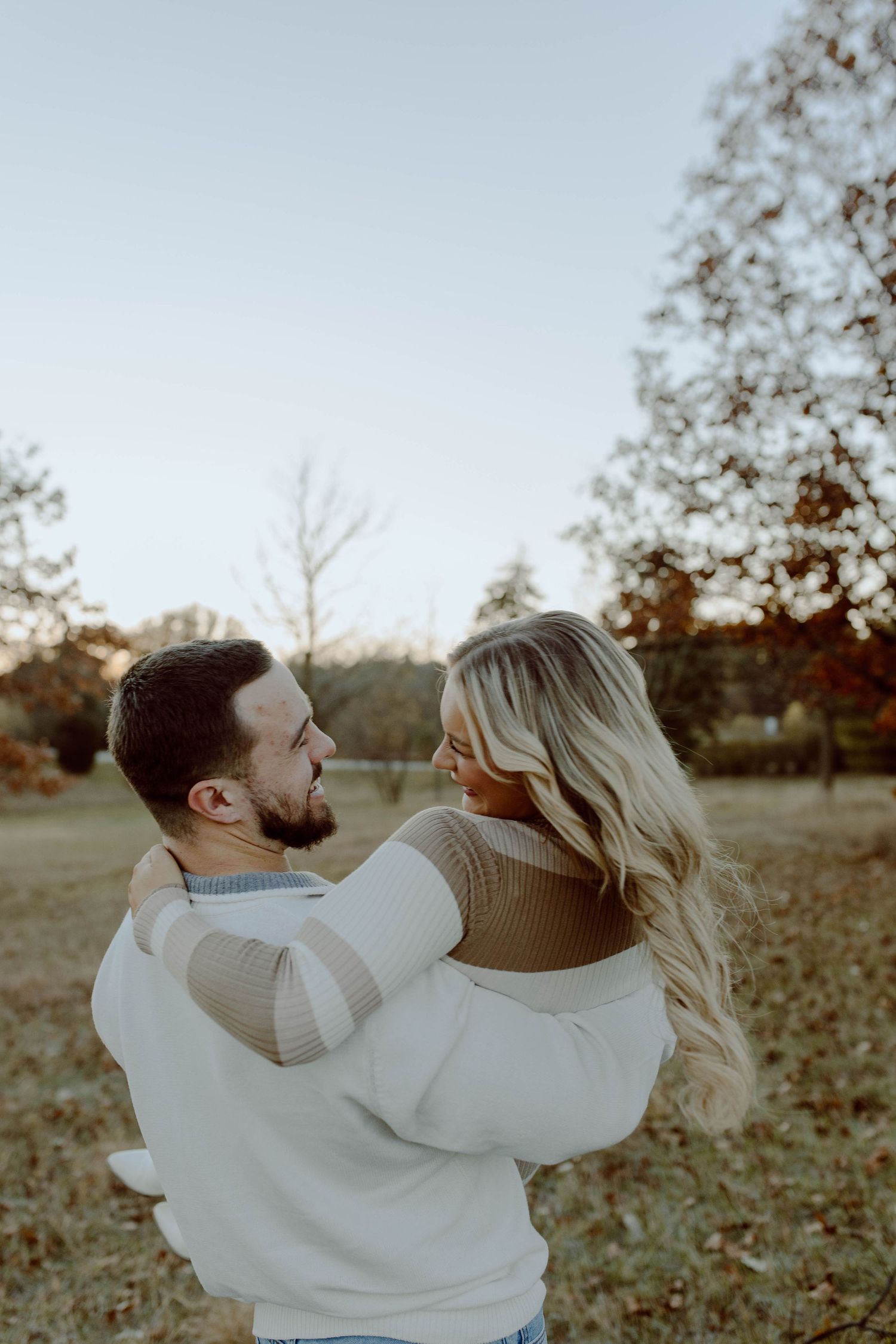 A couple in matching white sweaters embrace outdoors during golden hour in an autumn setting.