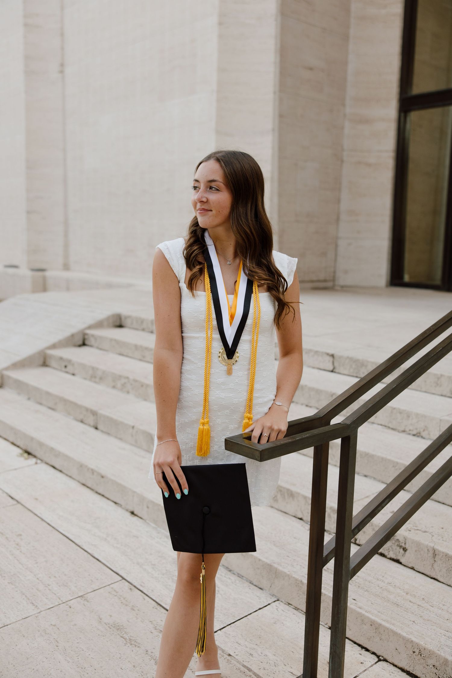 Graduate wearing academic regalia poses on steps with handrail at university.