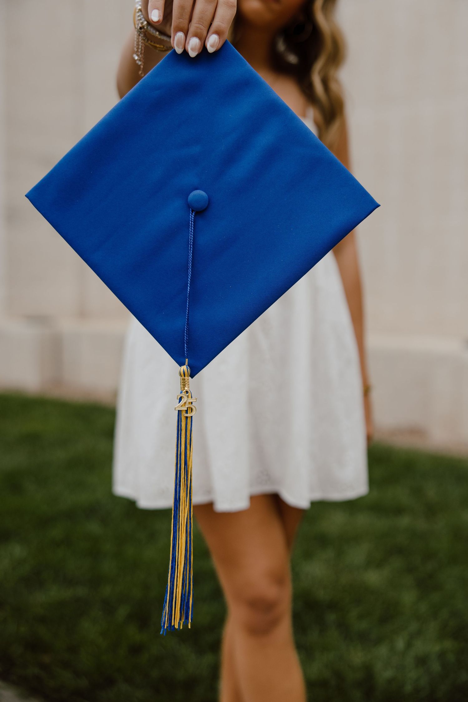 Blue graduation cap with gold tassel against white dress and grass background.