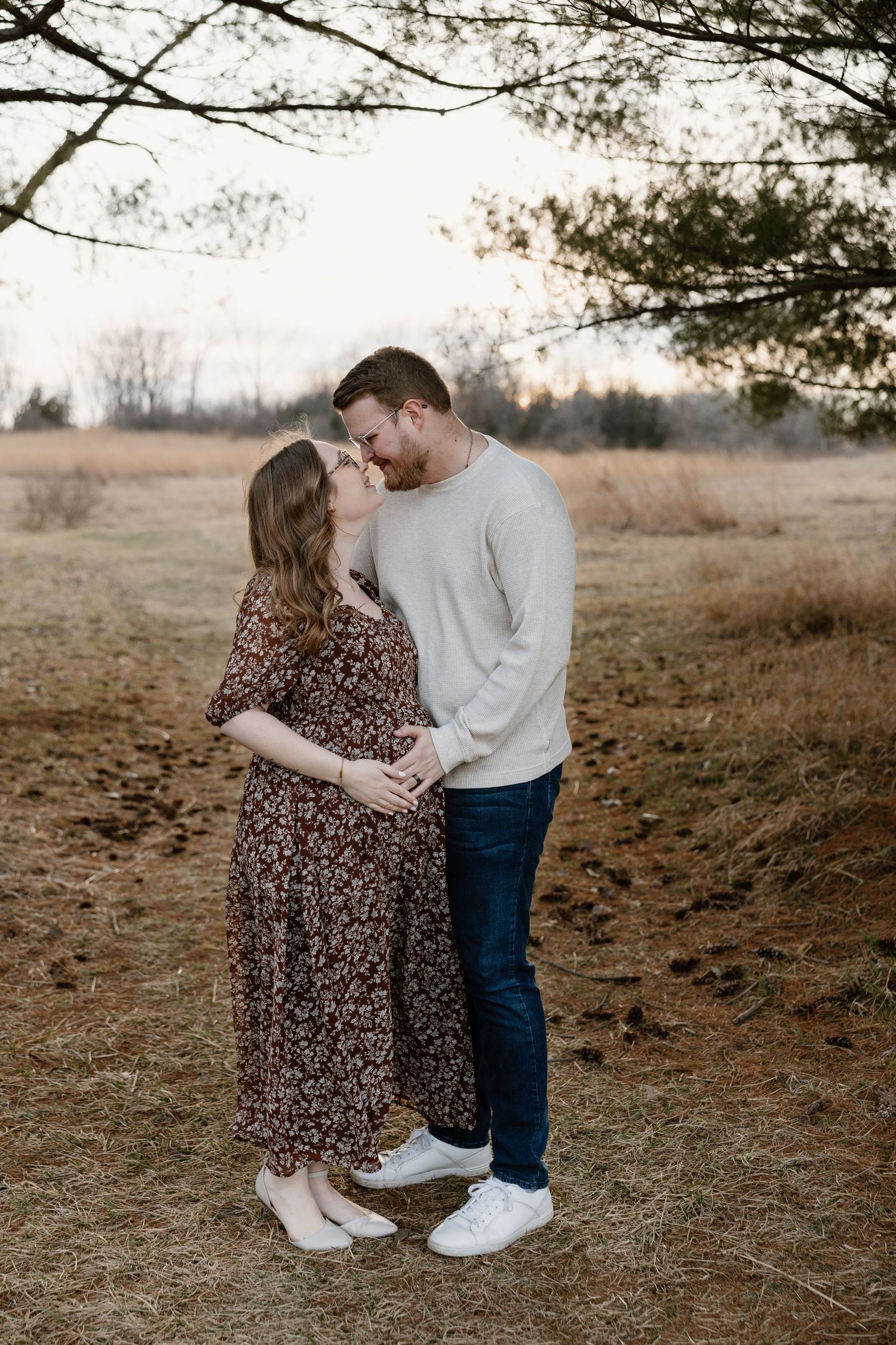 Pregnant couple sharing a tender kiss outdoors in a field at golden hour, woman in floral dress, man in cream sweater.