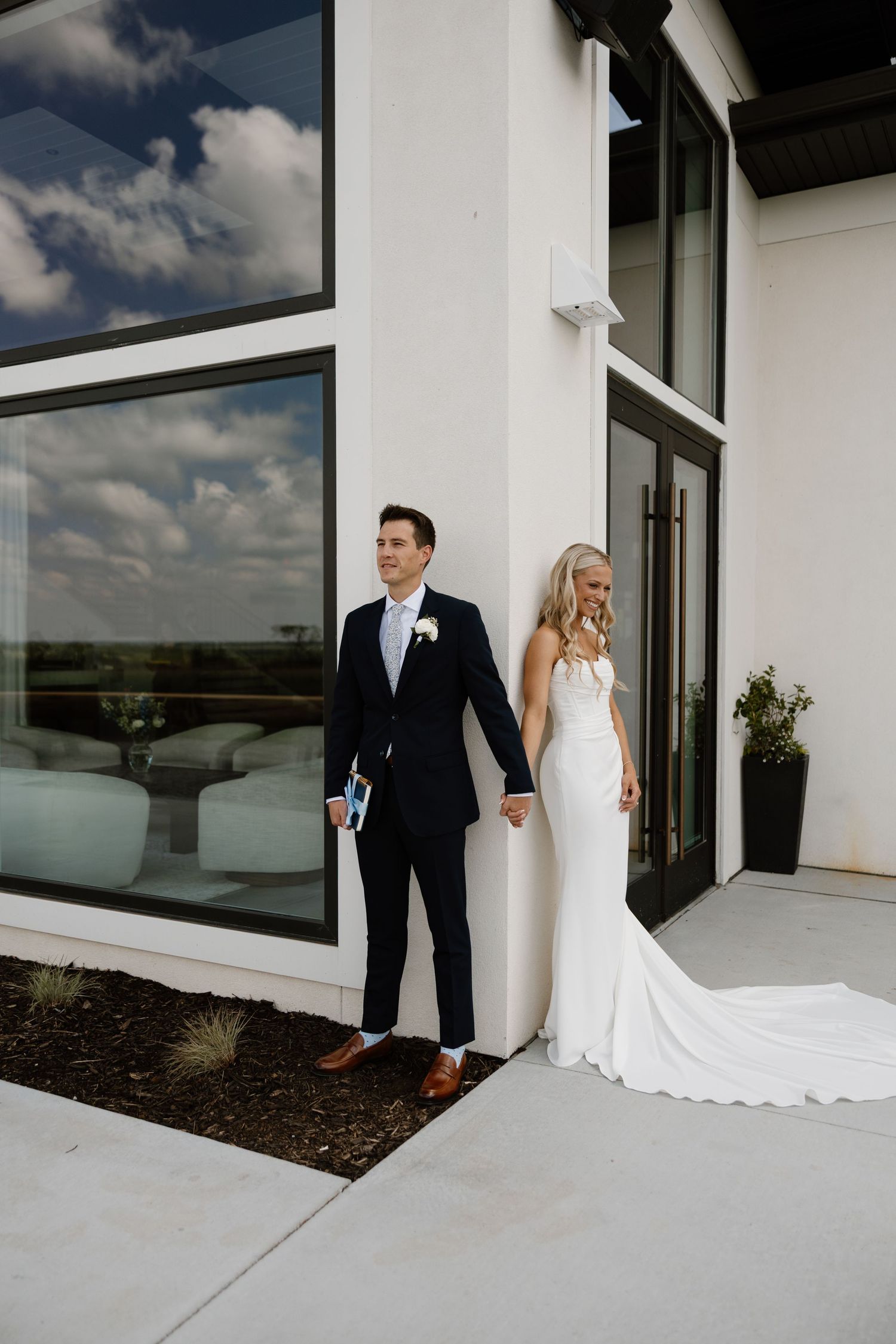 Bride in white gown and groom in navy suit pose outside modern white building on their wedding day.