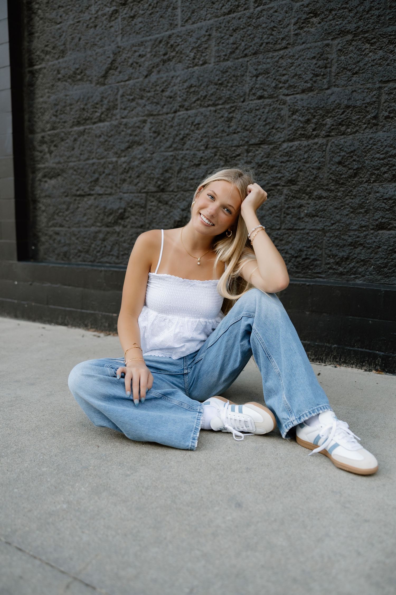 Relaxed poses against dark wall wearing white top, jeans and sneakers.