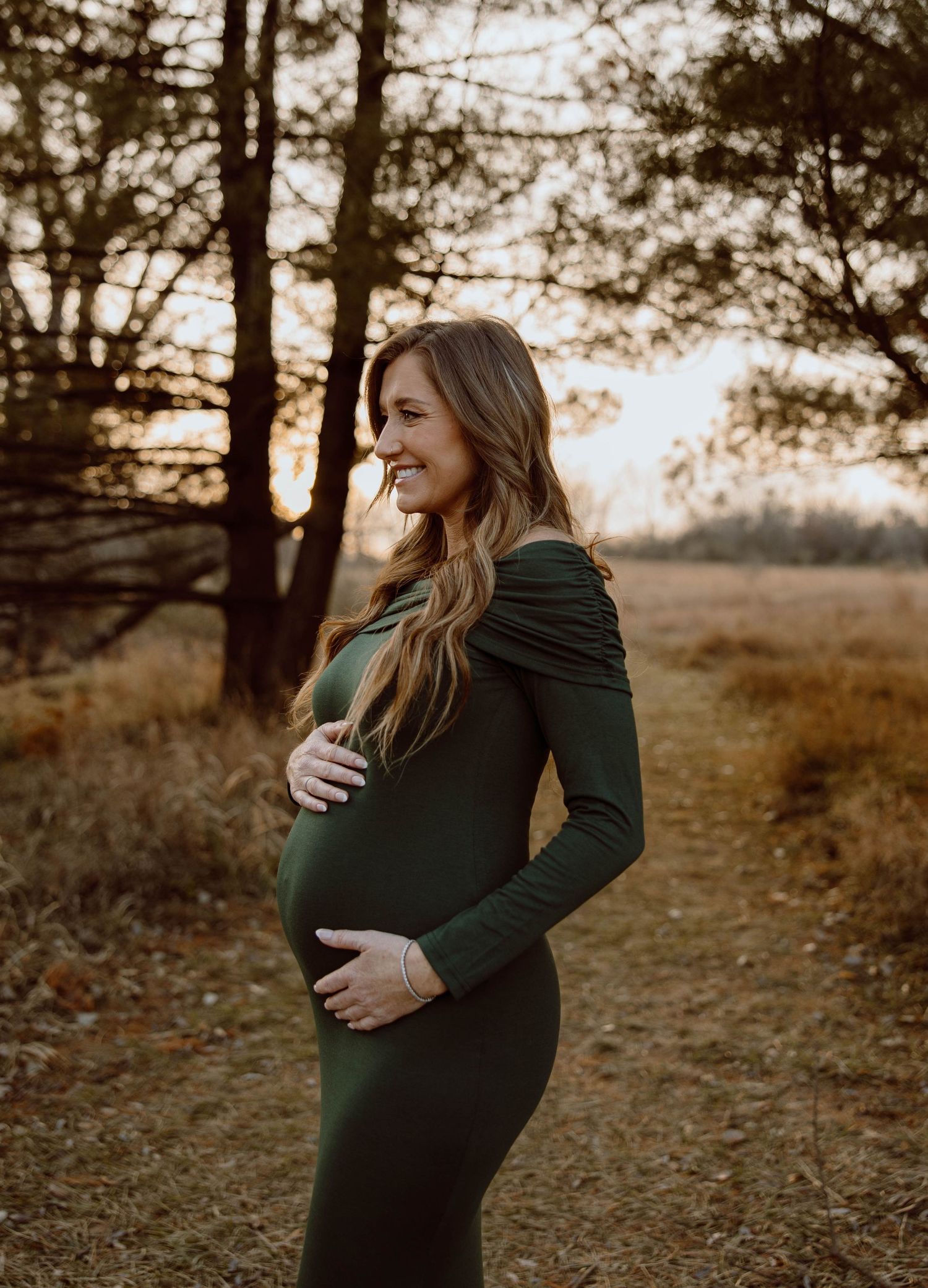 Maternity photo of woman in dark green dress at golden hour in forest setting.