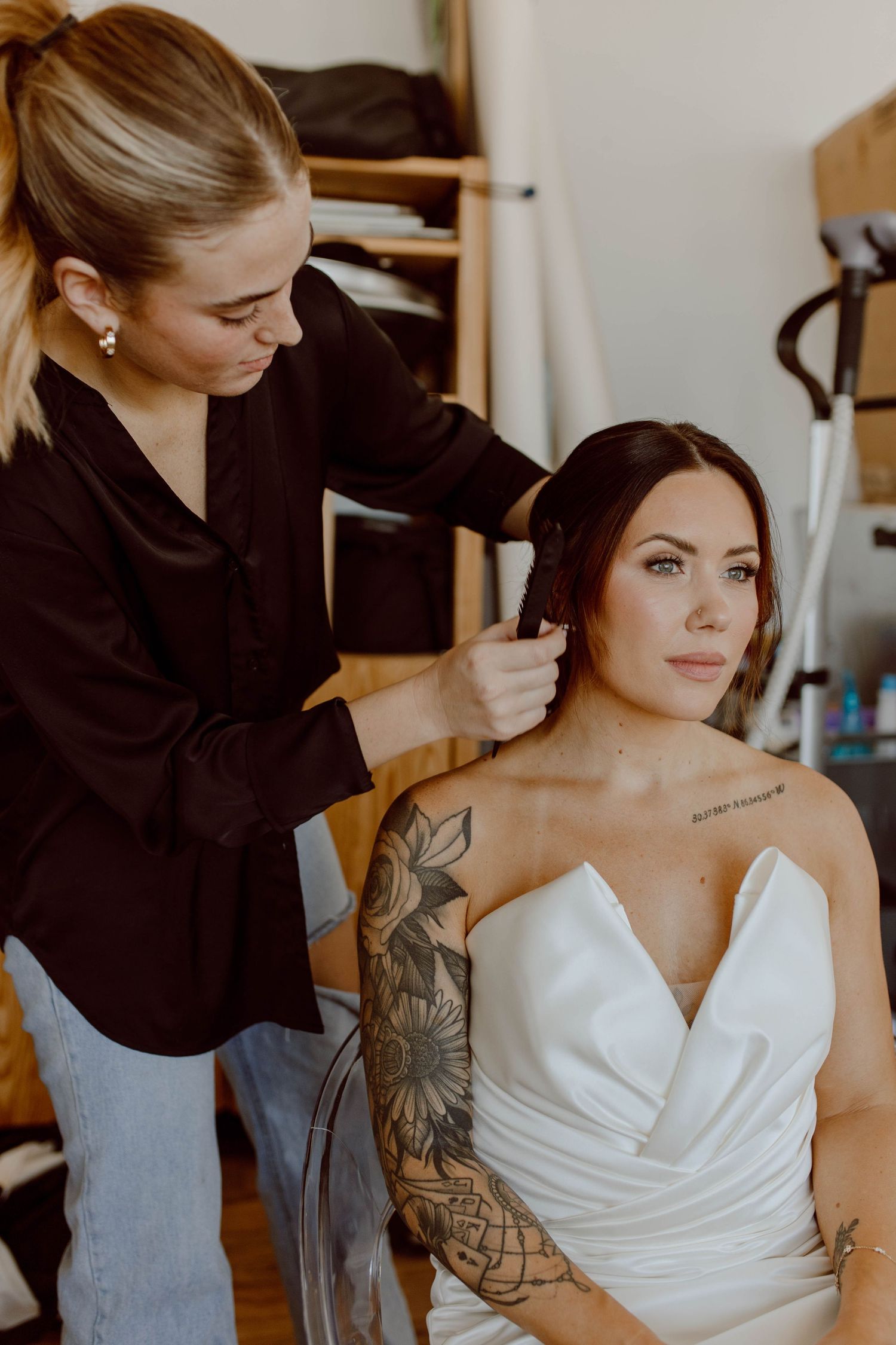 A makeup artist styles a bride's hair as she sits in a white wedding dress showing her floral arm tattoo.
