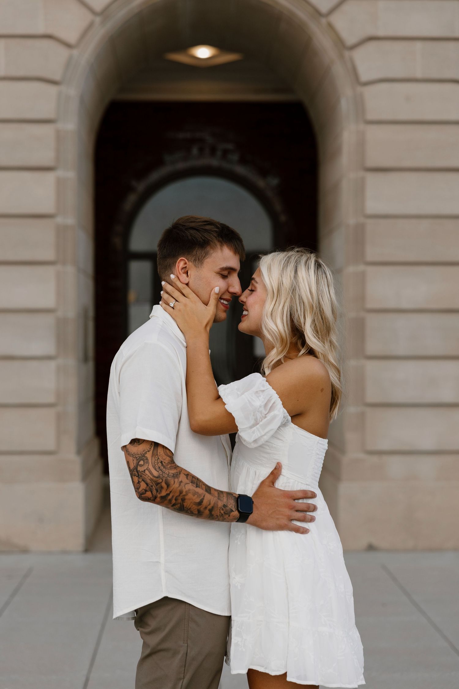 A couple embraces in front of arched doorways of a classical stone building.