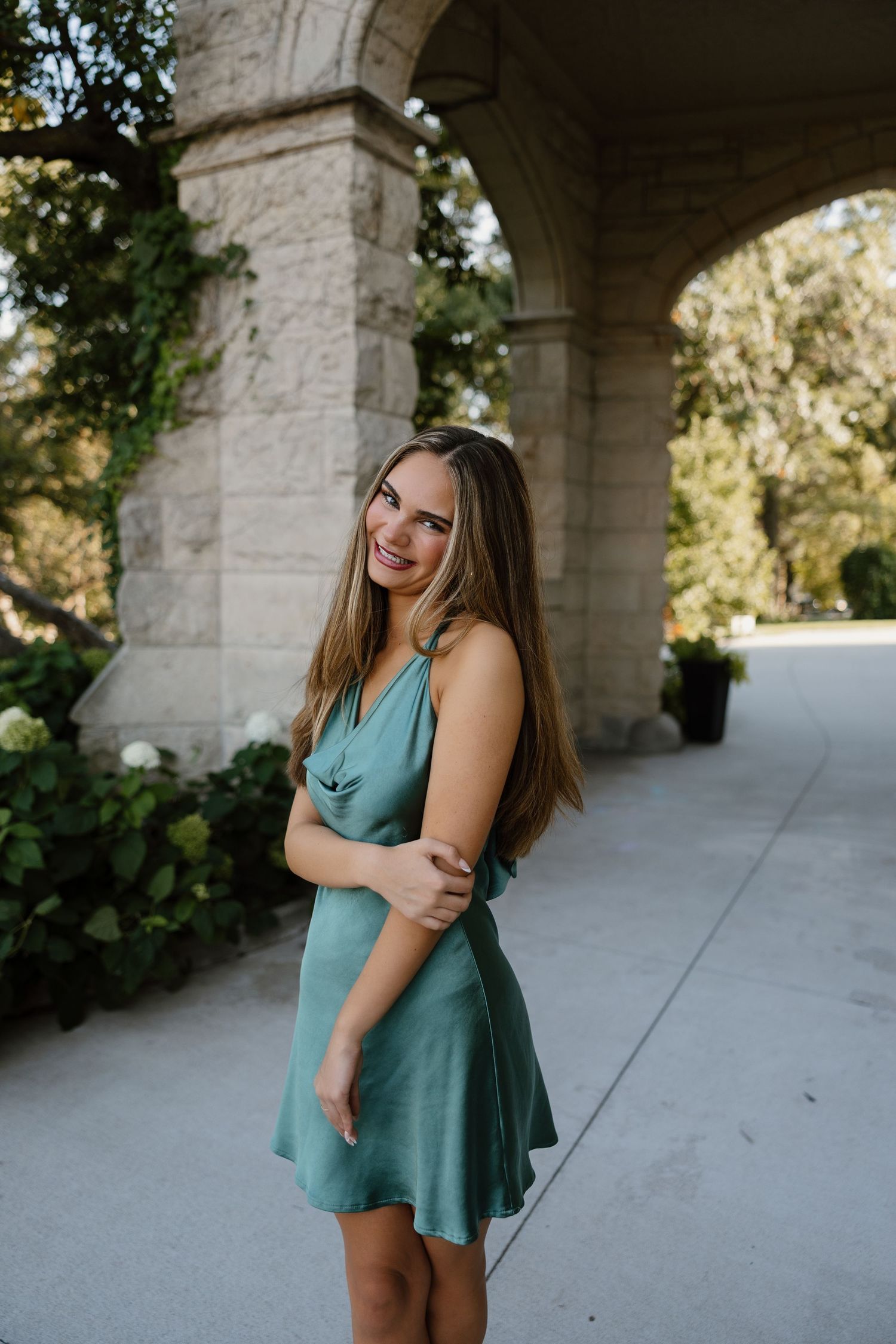 A woman in a teal dress poses outdoors under stone archways in a garden setting.