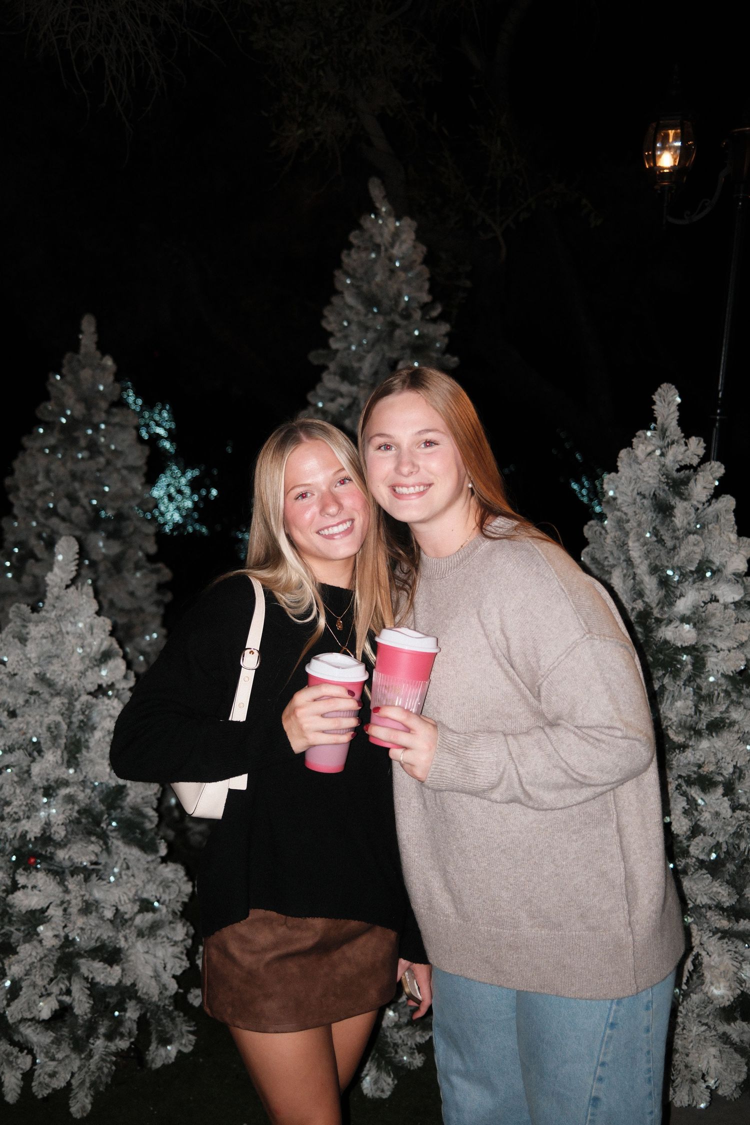 Two people holding red holiday drinks stand in front of snow-covered Christmas trees at night with decorative lighting.