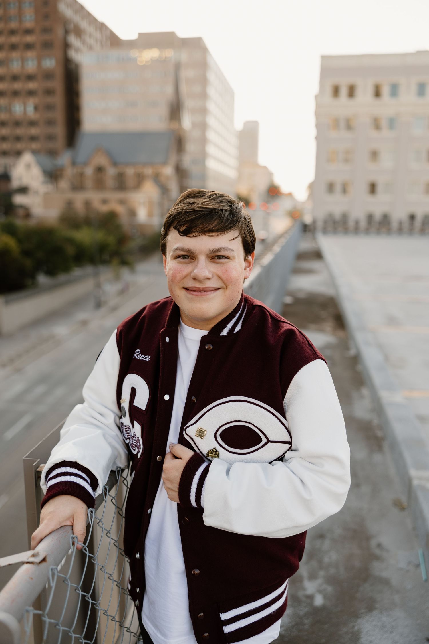 A student wearing a maroon and white varsity letterman jacket poses on a city bridge during golden hour.