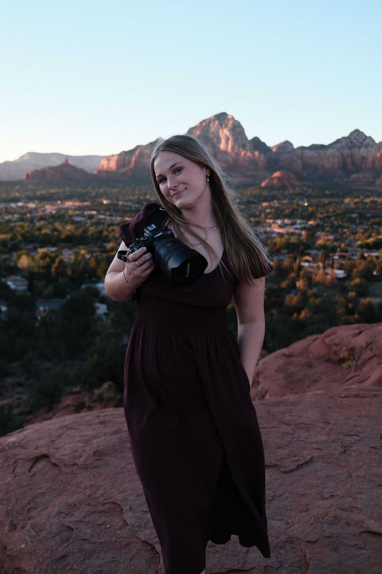 A photographer with camera stands on red rocks overlooking Sedona's mountain landscape at dusk.