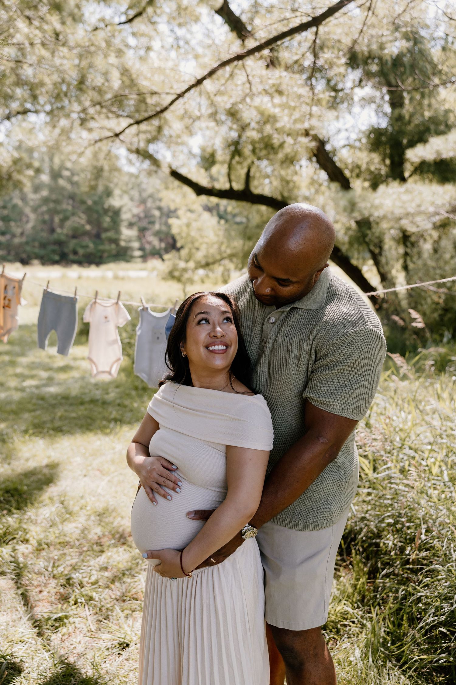 Expecting couple poses together in a sunny outdoor setting with baby clothes hanging on a clothesline.