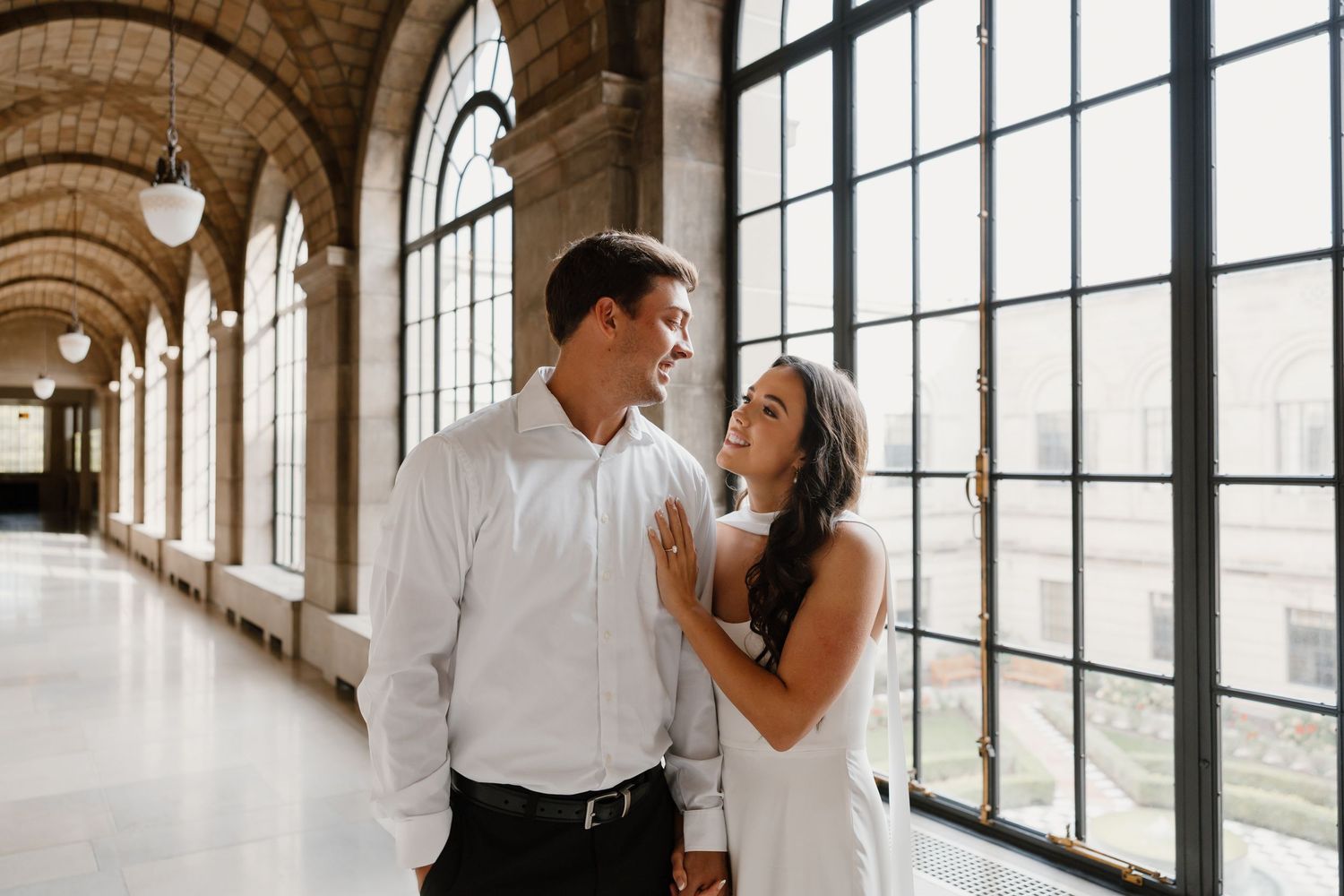 Couple shares intimate moment in elegant arched hallway with large windows.
