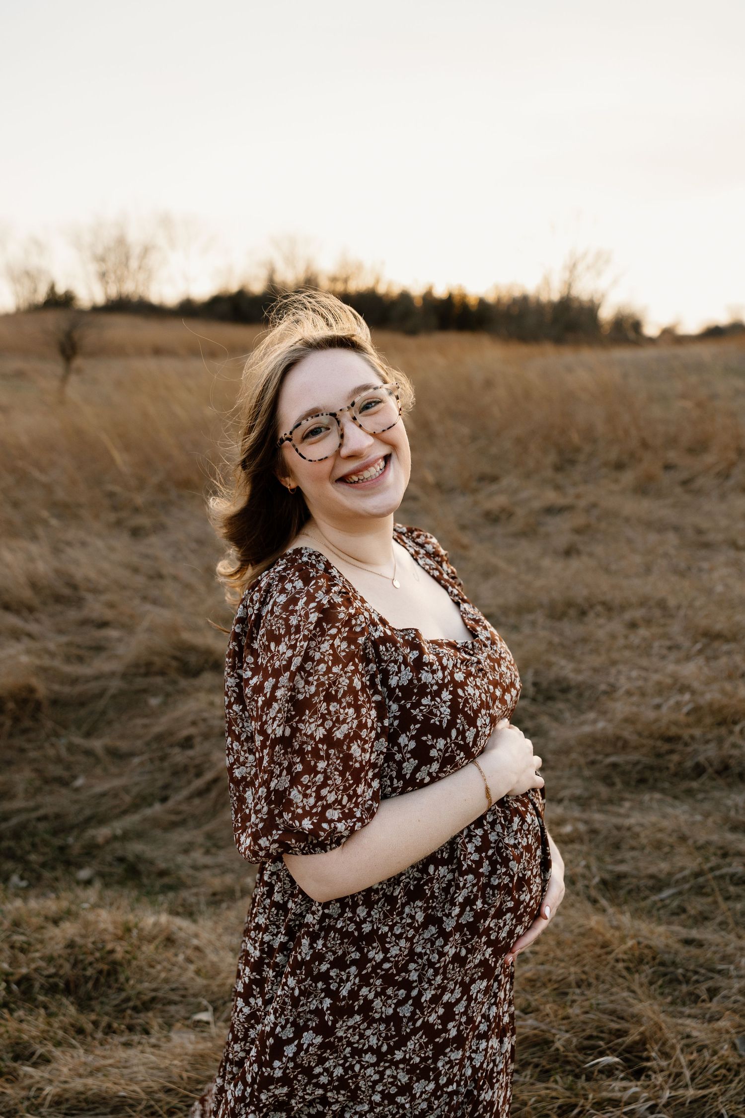 Smiling pregnant woman in floral dress and glasses standing in golden field at sunset.