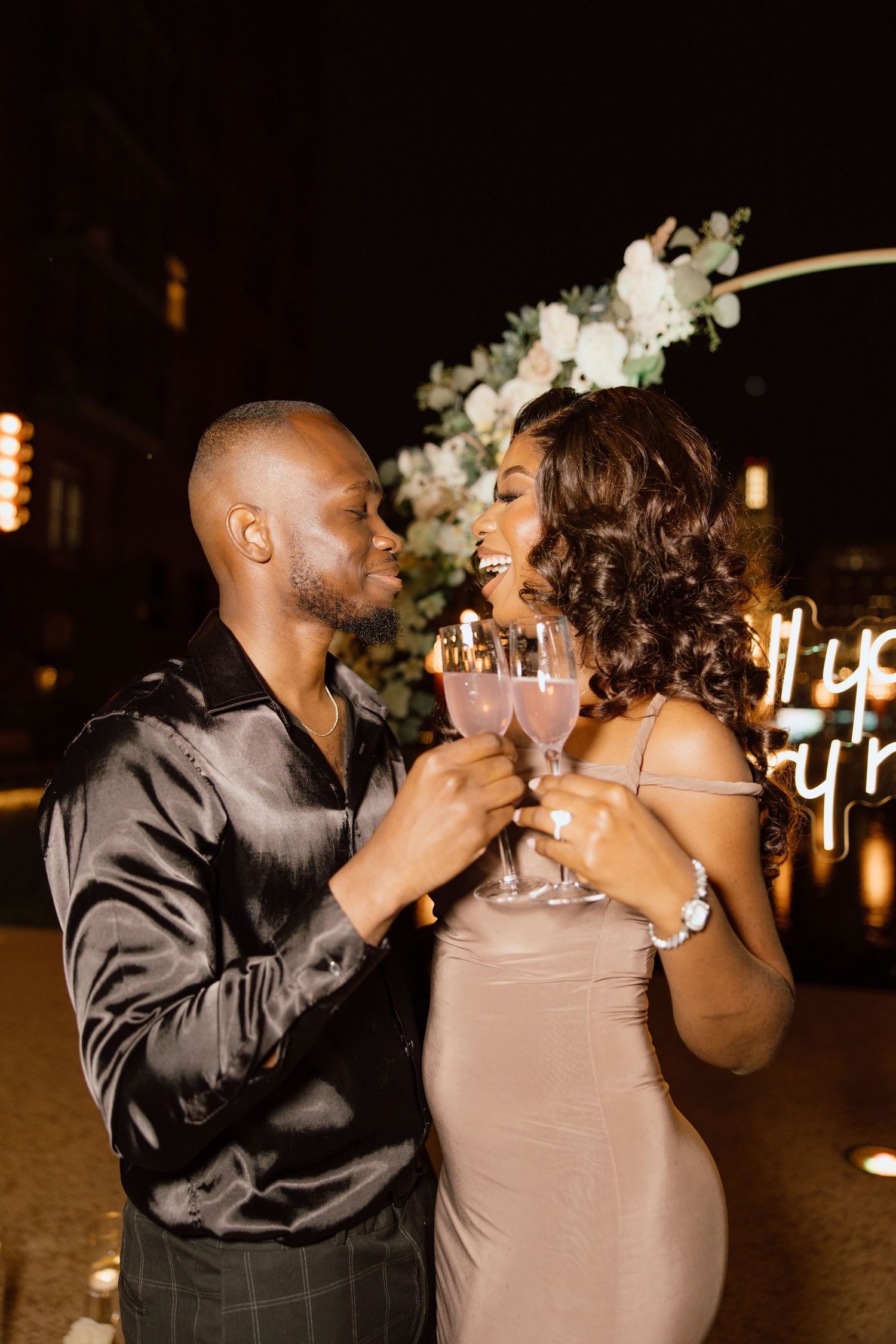 Couple celebrates engagement with champagne toast under evening lights.