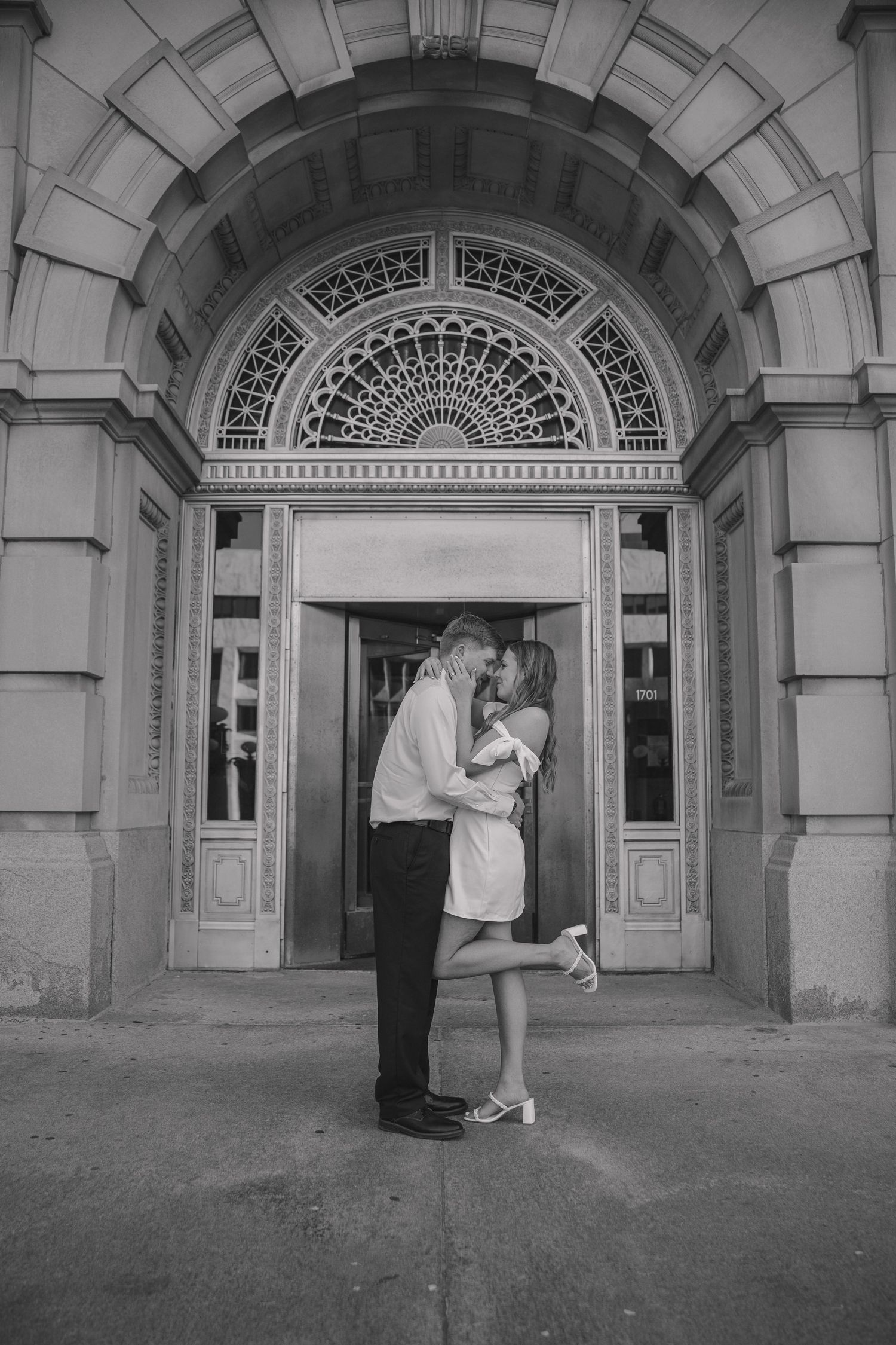 A black and white photo of a couple sharing a tender moment under an ornate architectural archway.