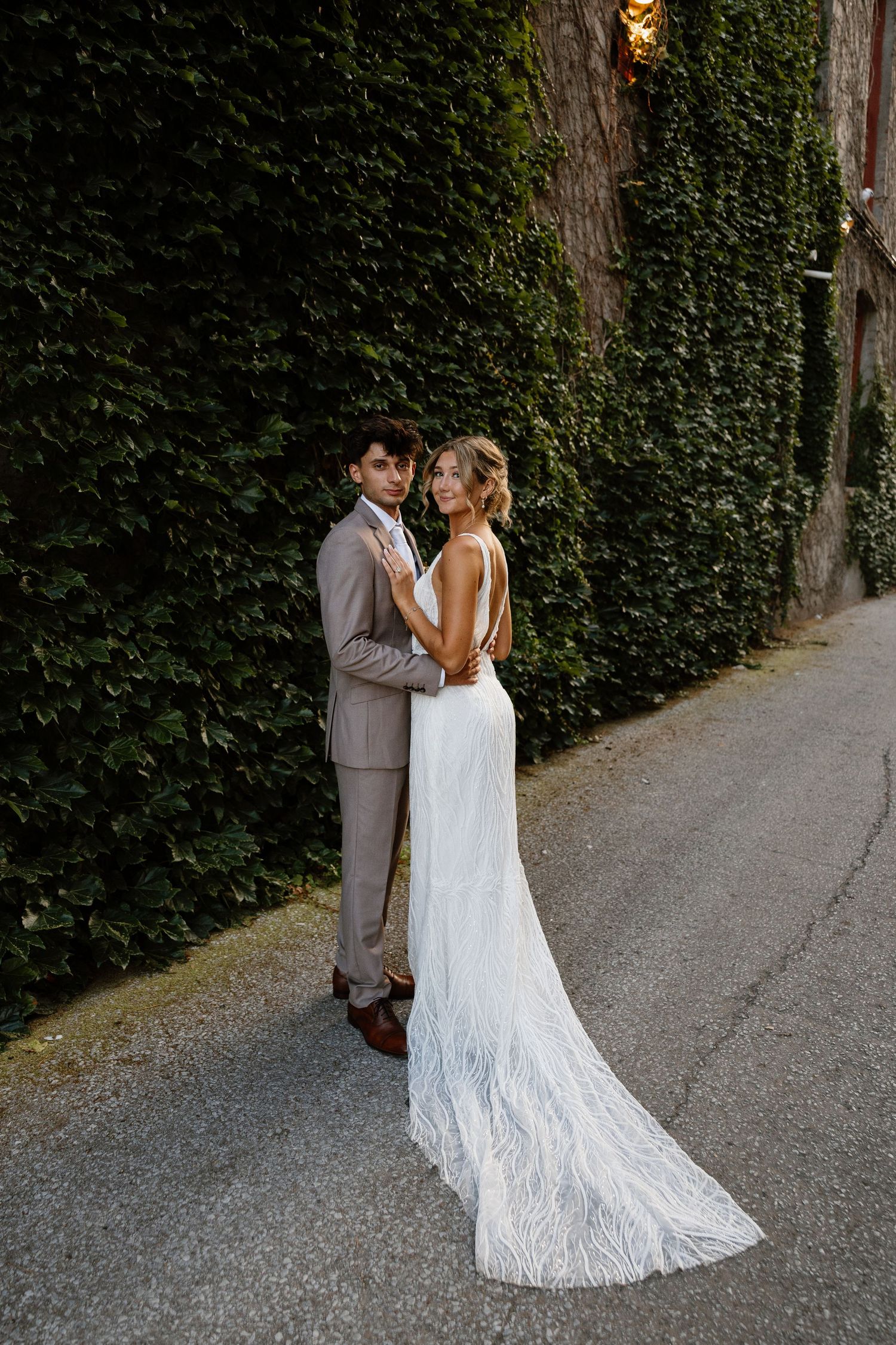 Bride in lace gown and groom in grey suit embrace on a pathway with ivy walls.