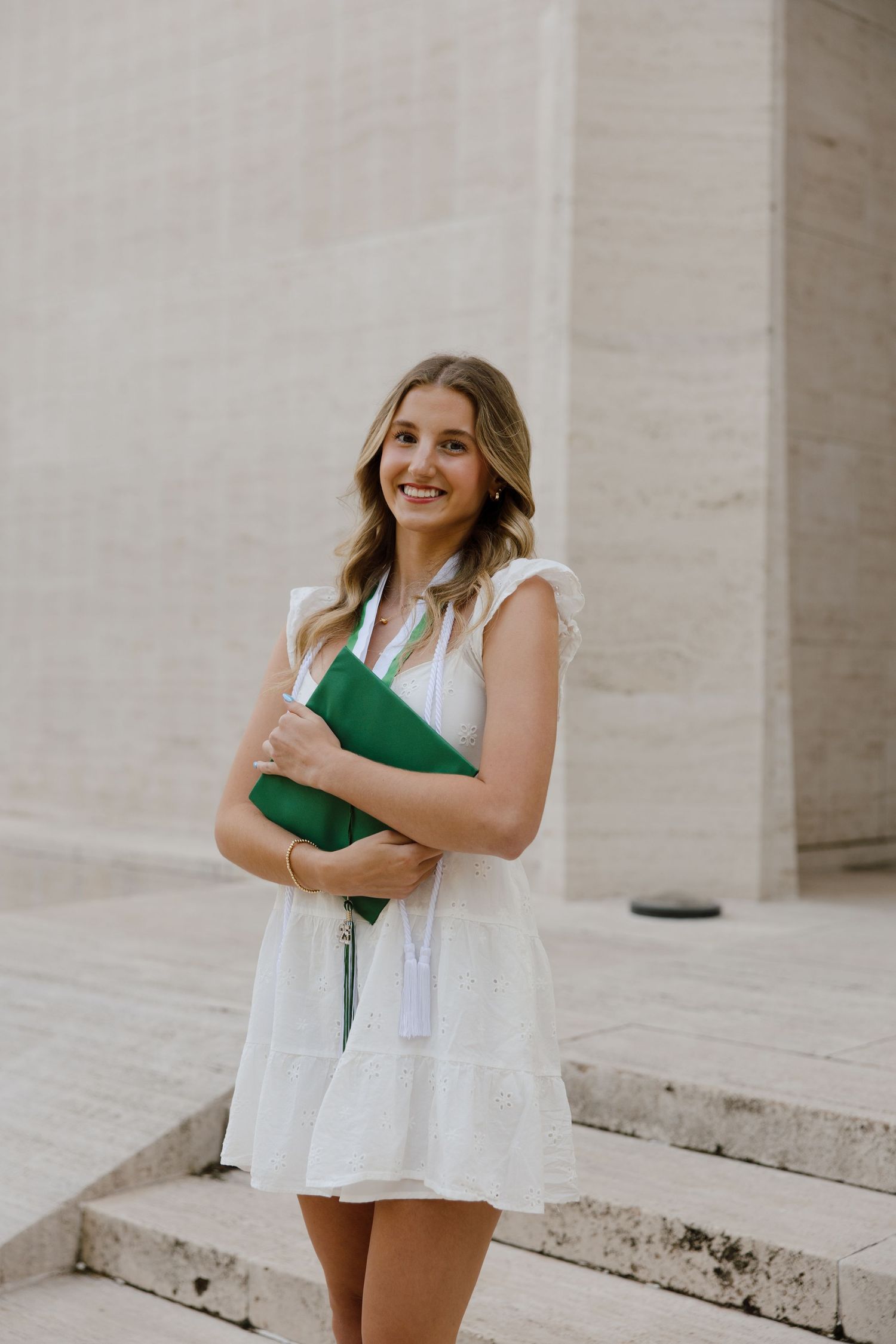 A woman in a white dress holds a green book while standing on concrete steps.