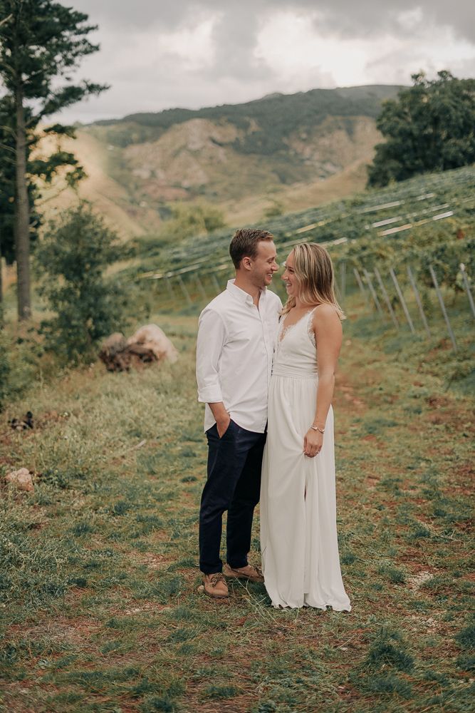 A romantic couple stands together on a hillside vineyard with mountains in the background on a cloudy day.
