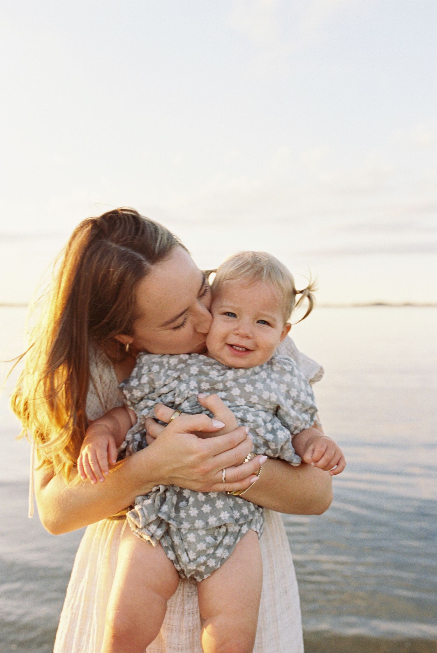 Parent and toddler sharing joyful cuddles in white dress against ocean backdrop.