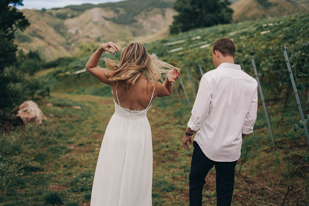 A couple in wedding attire walks through a scenic mountain landscape with rolling green hills in the background.