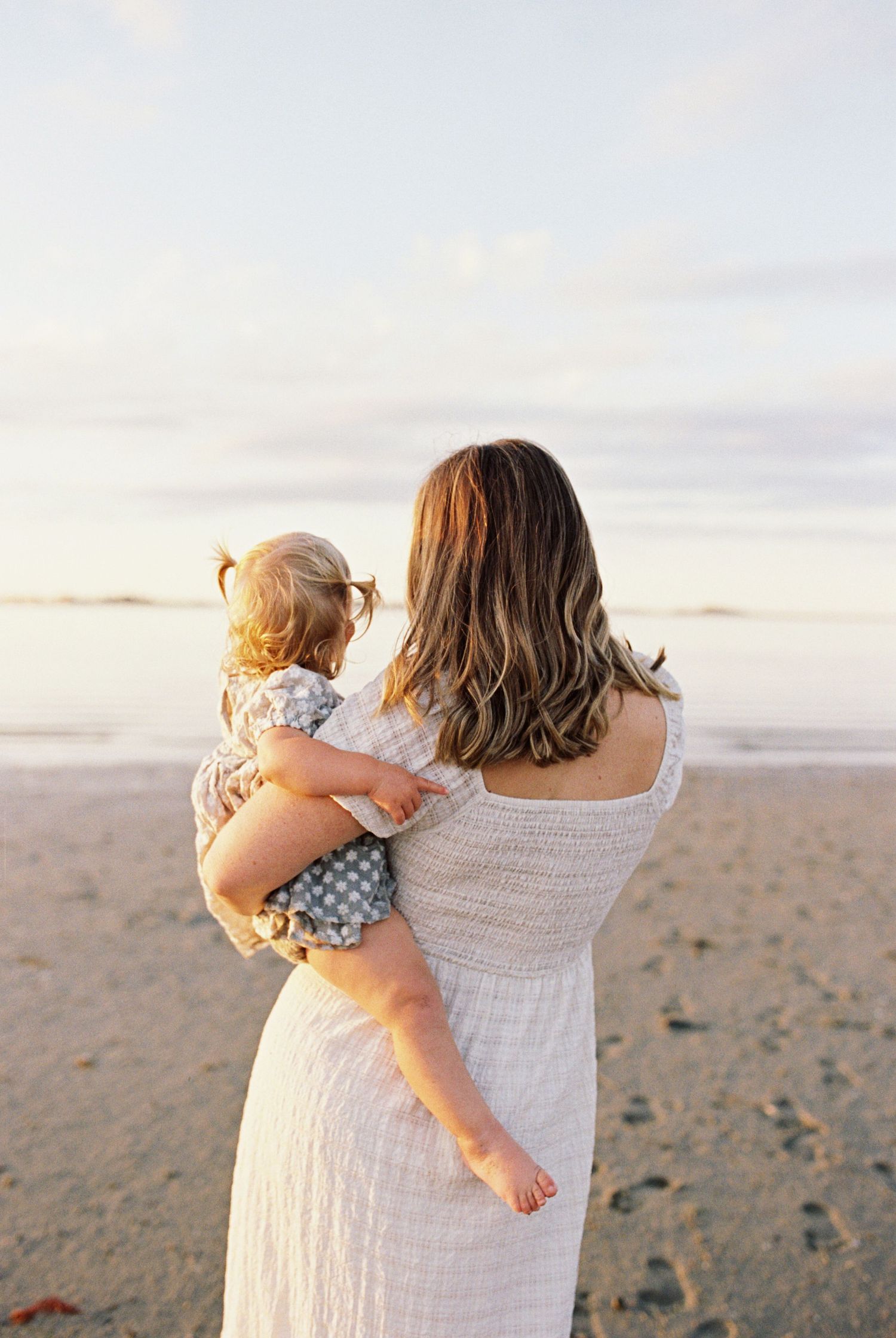Adult carrying toddler while walking along sandy beach at dusk.