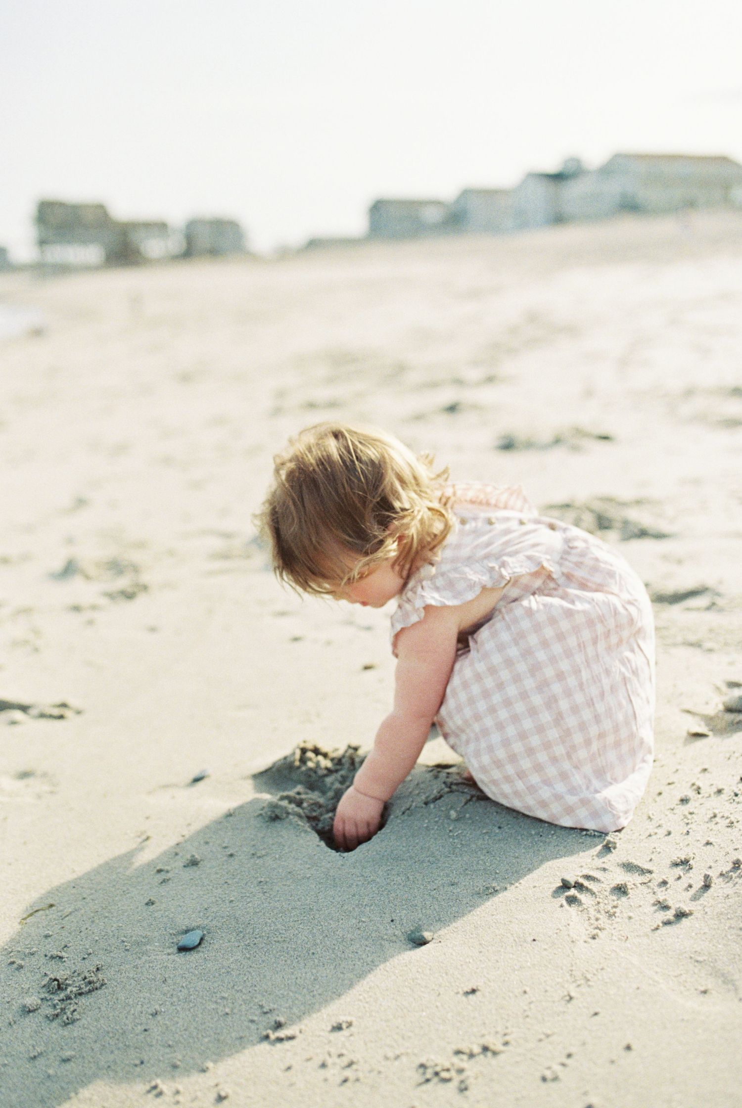 Small child in checkered dress exploring sand on sunny beach.