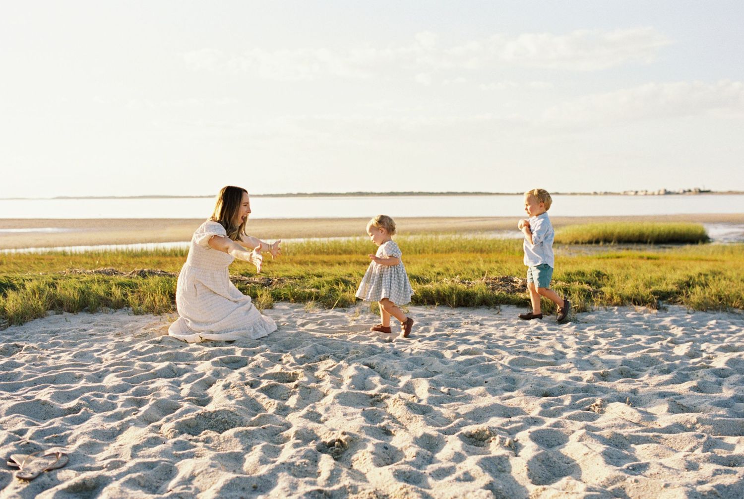 People enjoy a sunny evening together on a sandy beach with marsh grass and water in the background.
