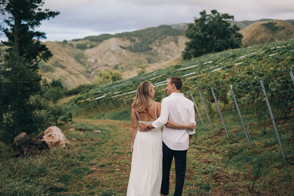 Two people in white outfits walk hand in hand through a scenic mountain landscape with green rolling hills.