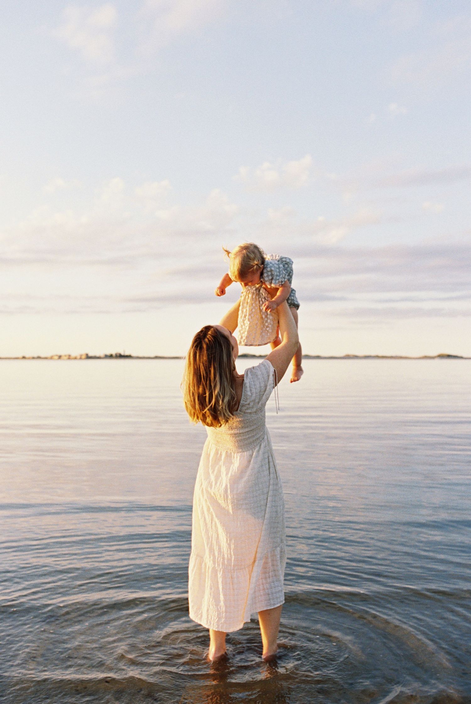 Adult in white dress lifting child playfully above water at beach during golden hour.