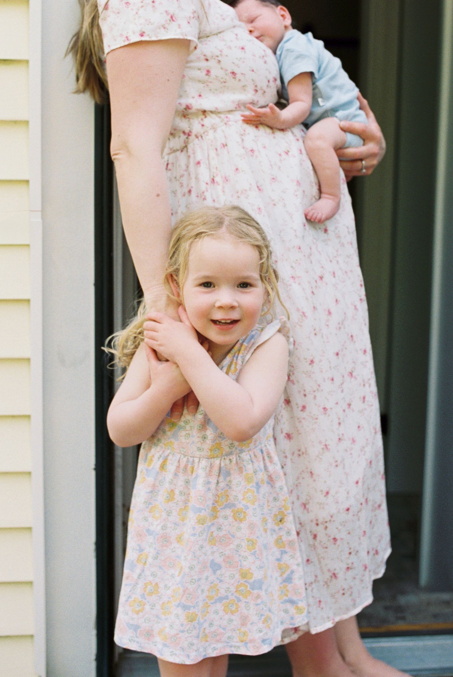 Young child in floral dress stands by family member holding sleeping baby.