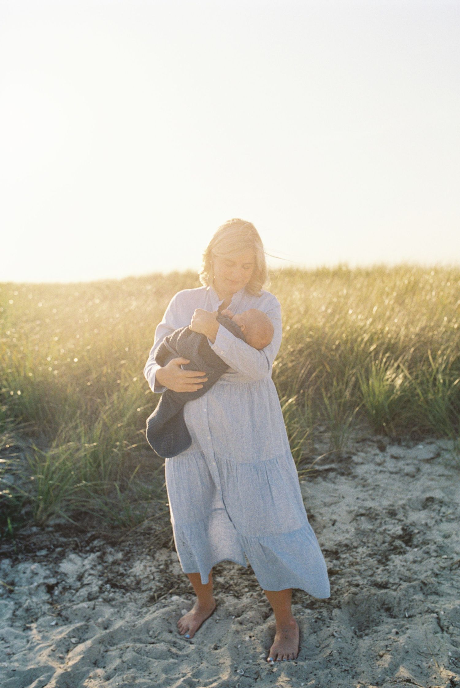 A person in a flowing white dress holds something close while standing on a beach at sunset.