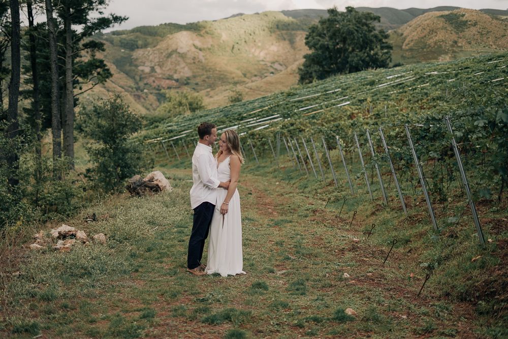 A couple in formal attire embraces on a scenic hillside path surrounded by lush greenery and rolling terrain.