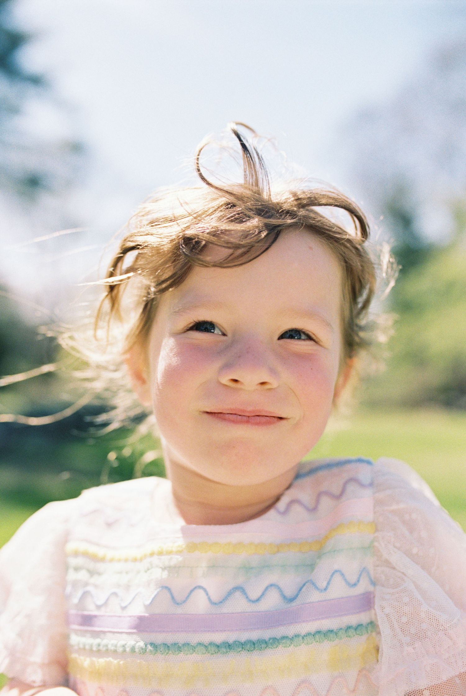 A young child smiles at the camera wearing a pastel striped shirt on a sunny day.