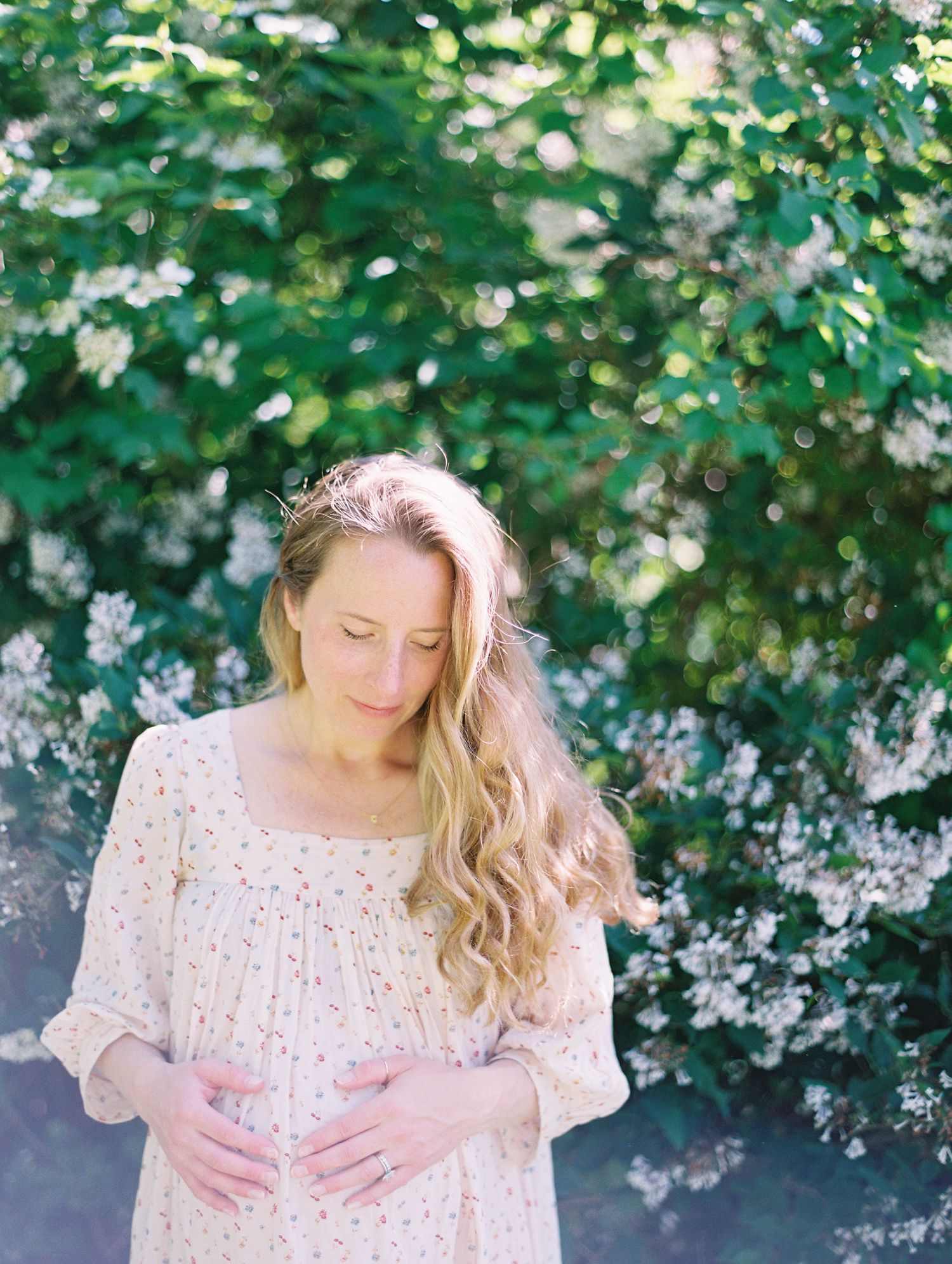 Woman in floral dress poses in garden with blooming white flowers.