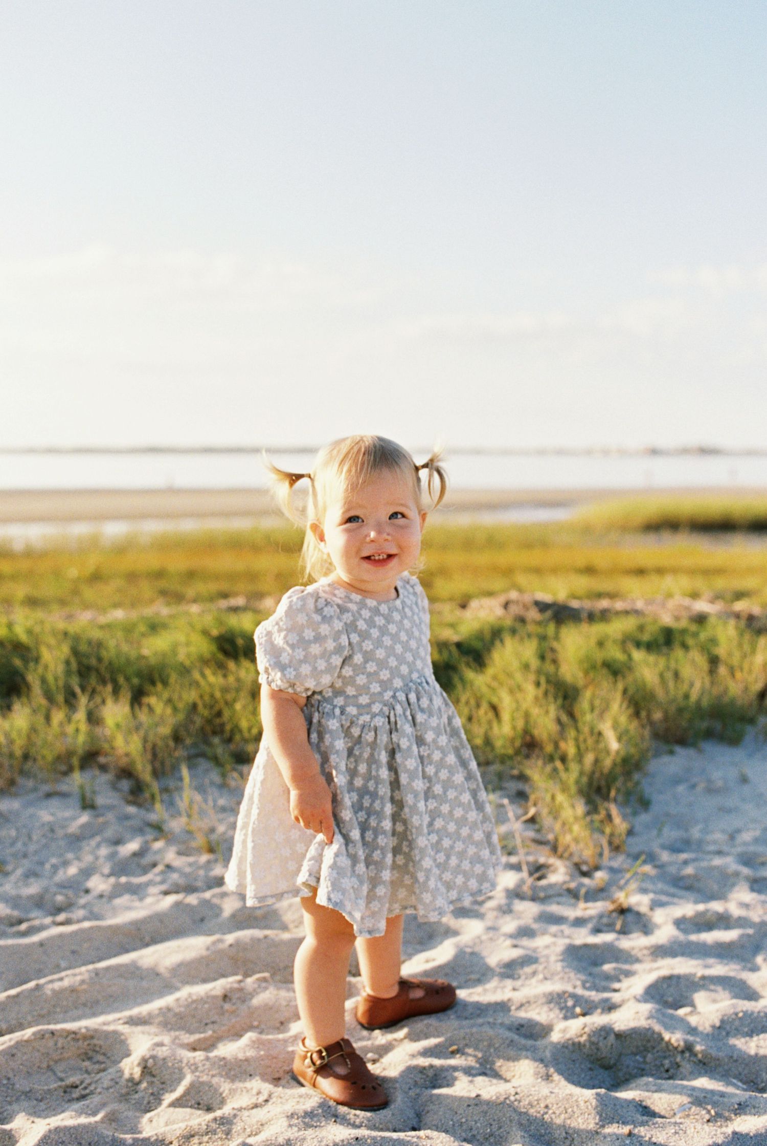 Toddler in polka dot dress playing happily in beach sand during sunset.