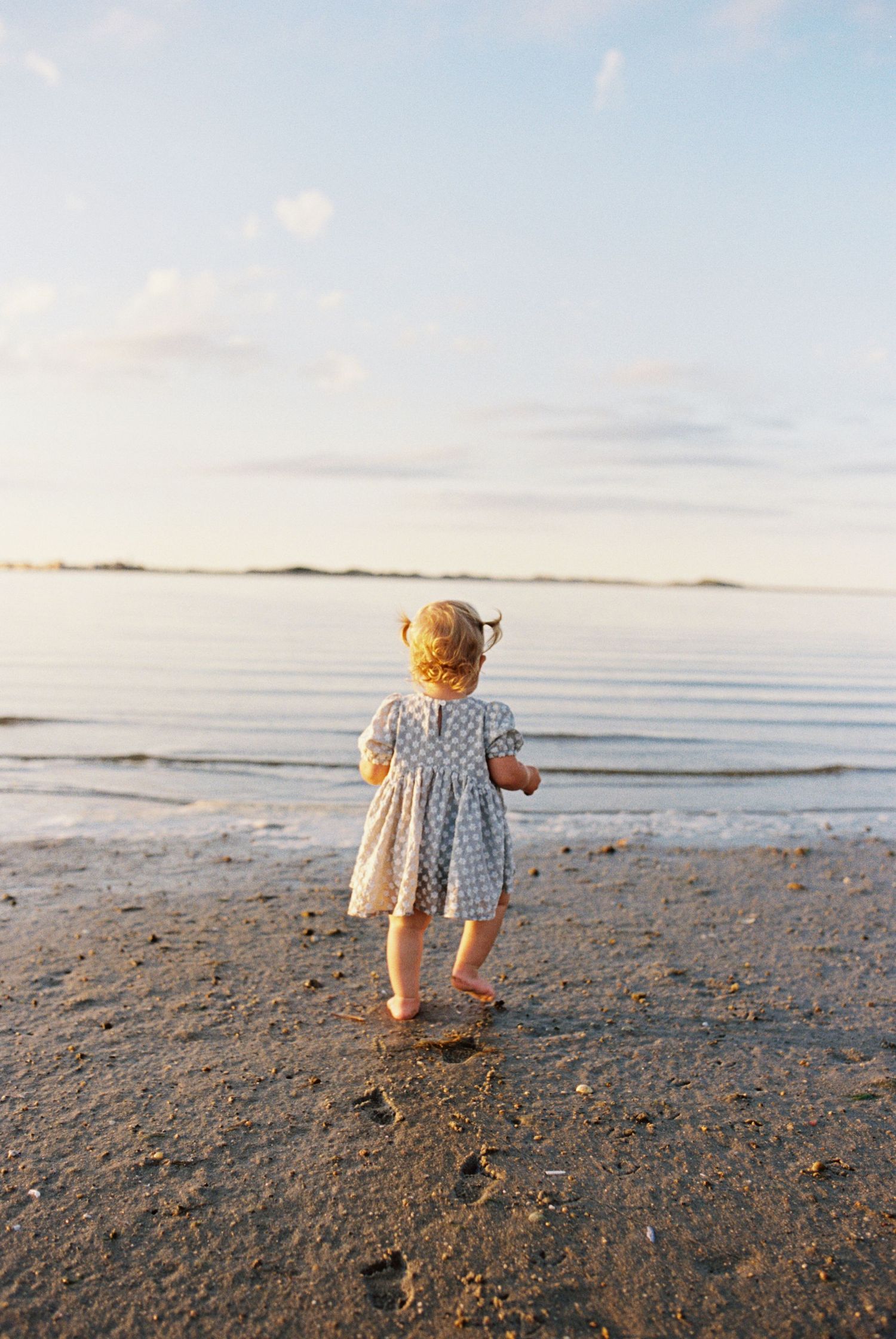 A small child in a patterned dress stands at the edge of calm waters during sunset.