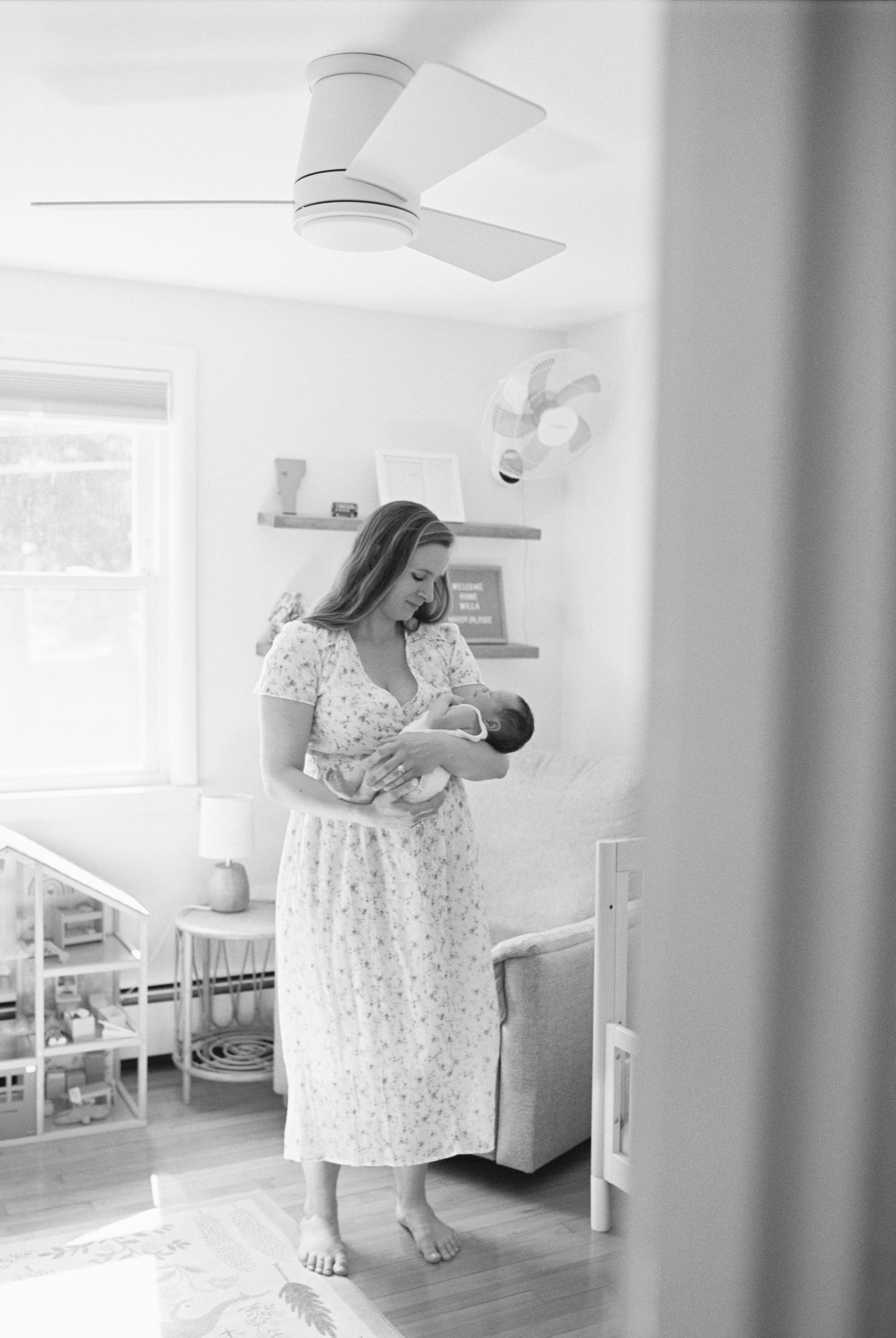 Mother in floral dress holding newborn in sunlit nursery.