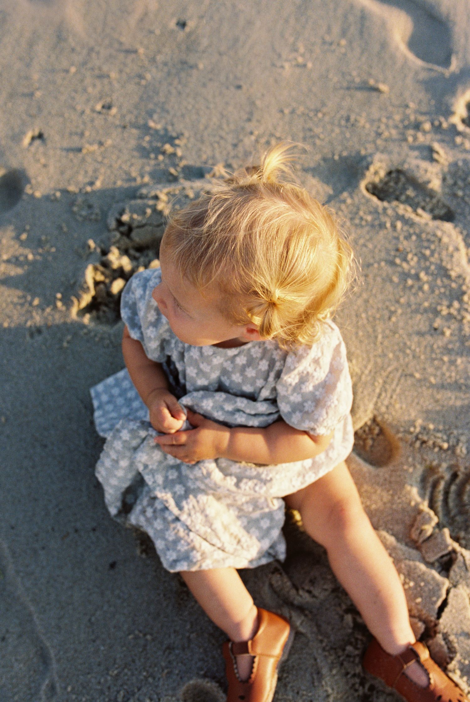 A child in a polka dot dress plays in the sand at the beach while wearing brown leather sandals.