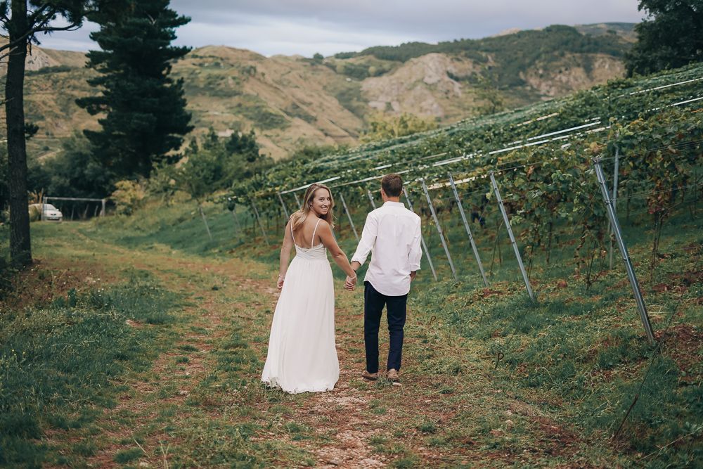 Couple walking hand in hand through scenic vineyard rows on rolling hills under cloudy sky.