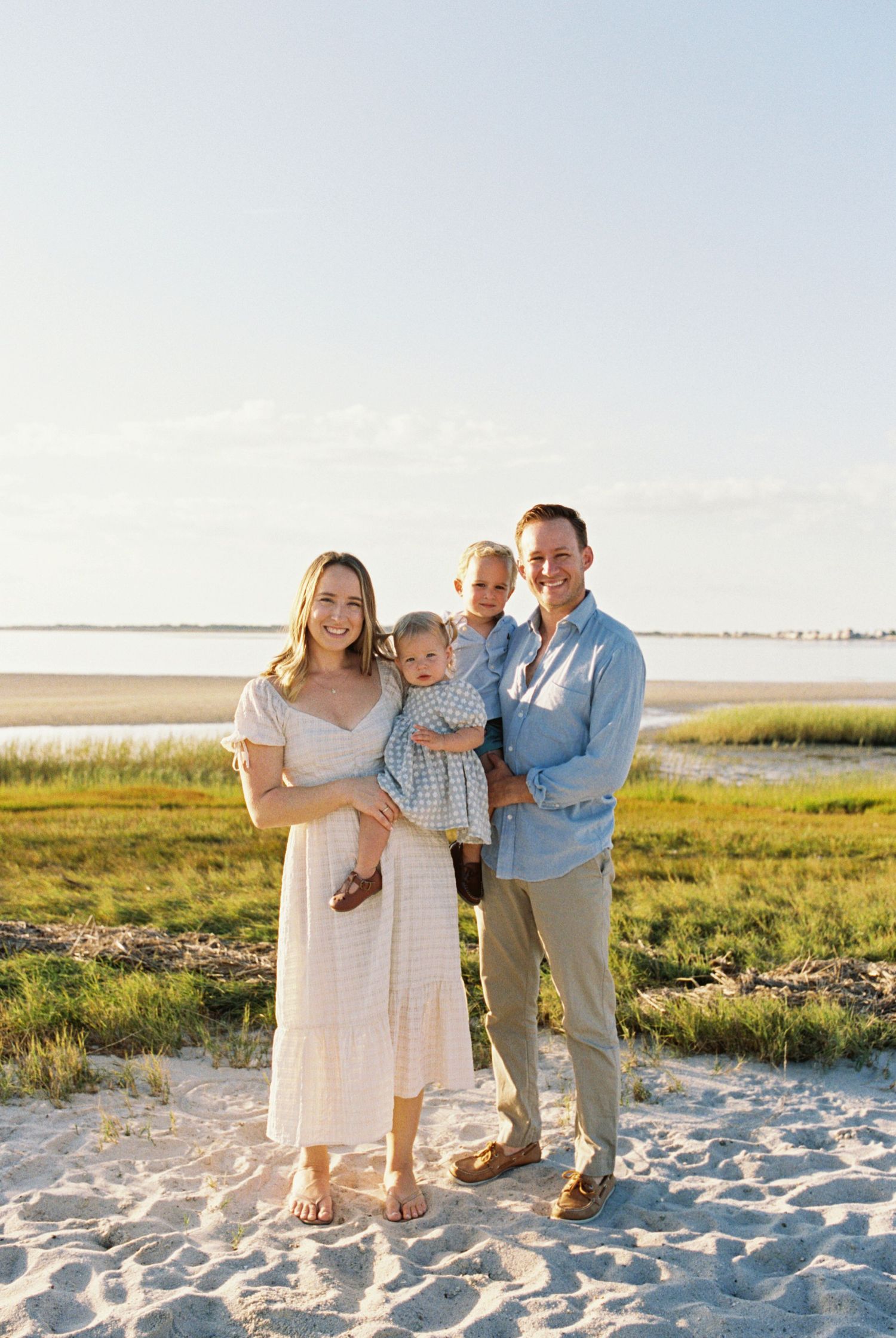 Family poses together on a sandy beach with marsh grass and water in the background at sunset.