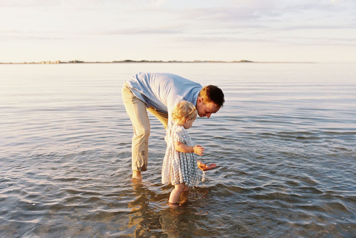 Person in white clothing bending down to explore shallow ocean waters at sunset.