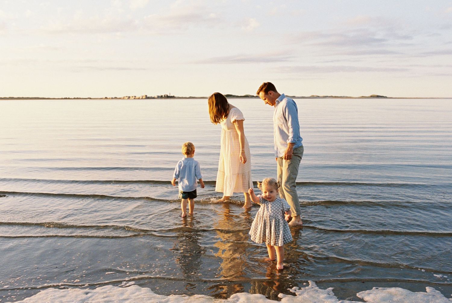 A serene beach scene at sunset shows people wading in calm waters along the shoreline.