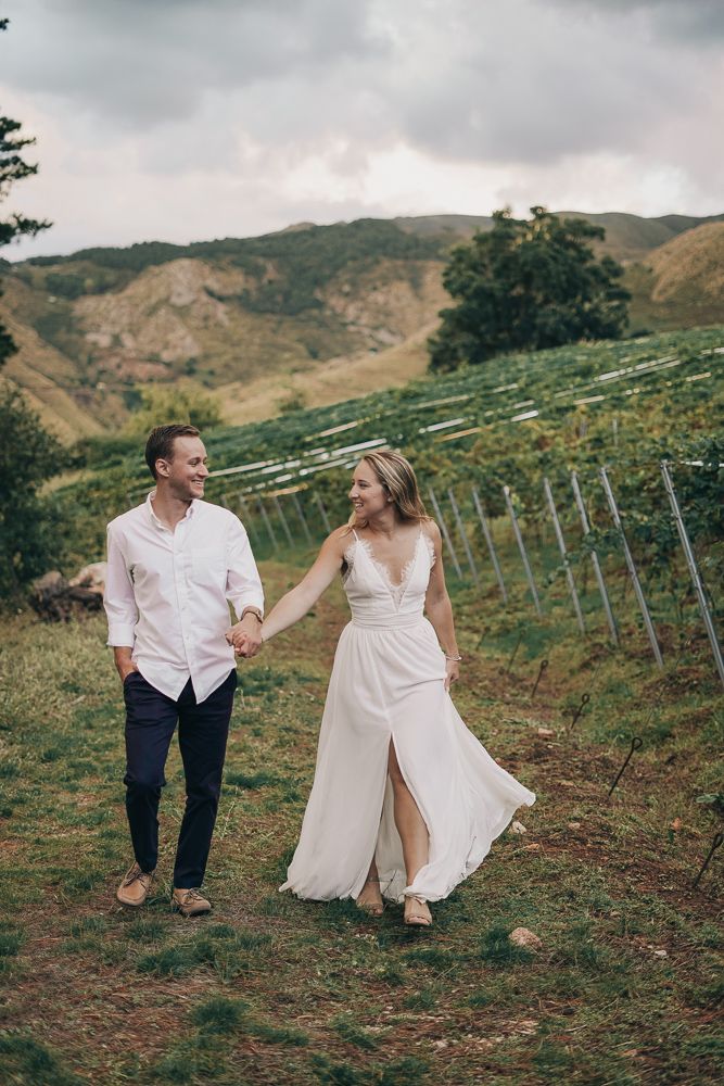 A couple in wedding attire walks through a scenic hillside vineyard on a cloudy day.
