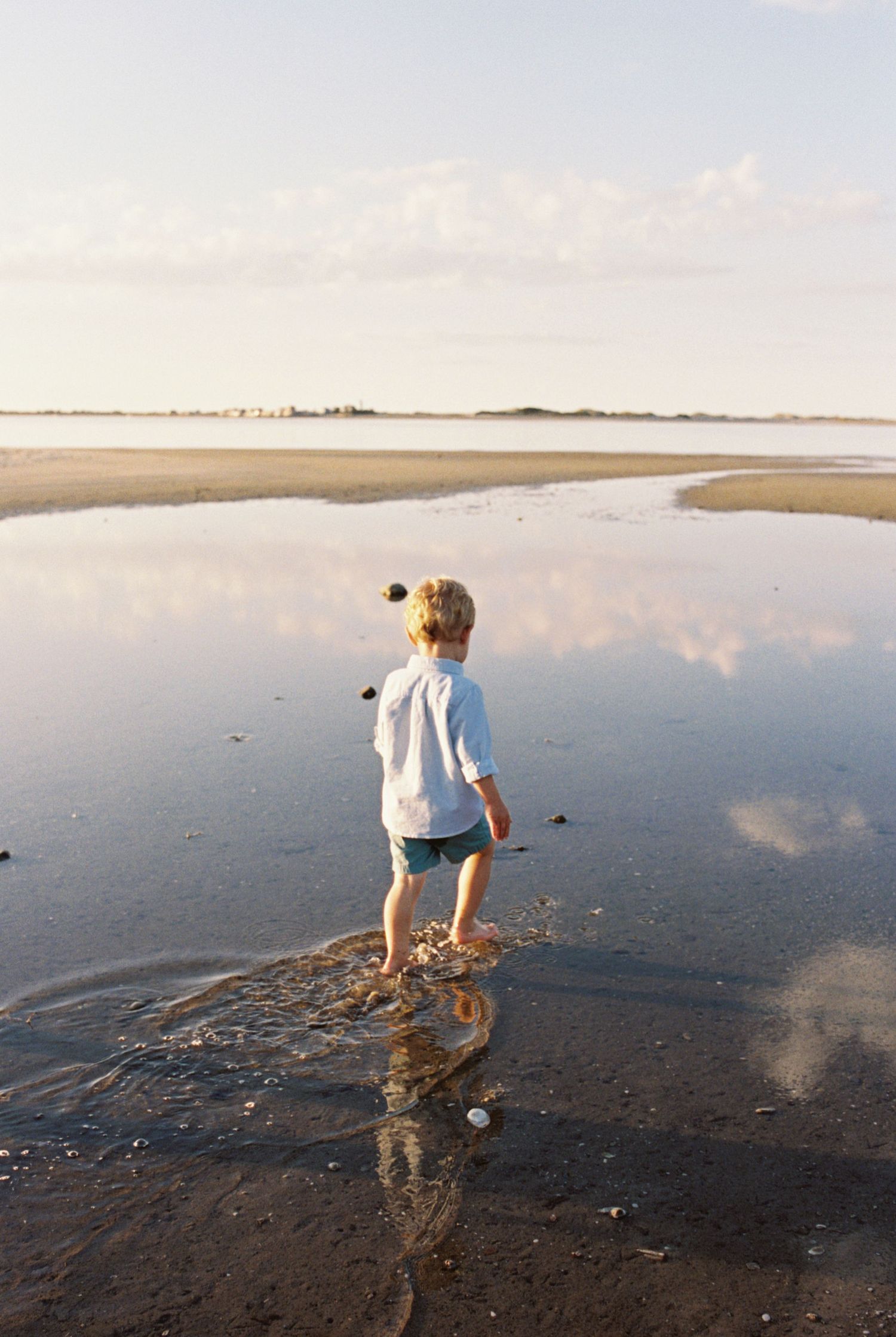 A person walks barefoot on a reflective wet beach at low tide during sunset, creating mirror-like reflections in the sand.