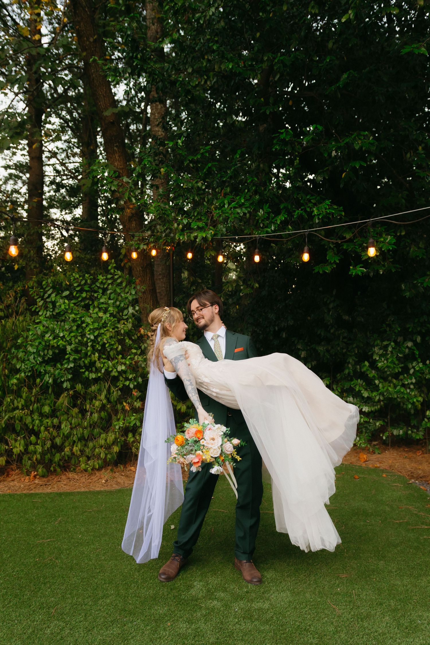 A couple performs romantic dip poses on a grassy lawn with string lights and trees in the background. Buford wedding photographer.