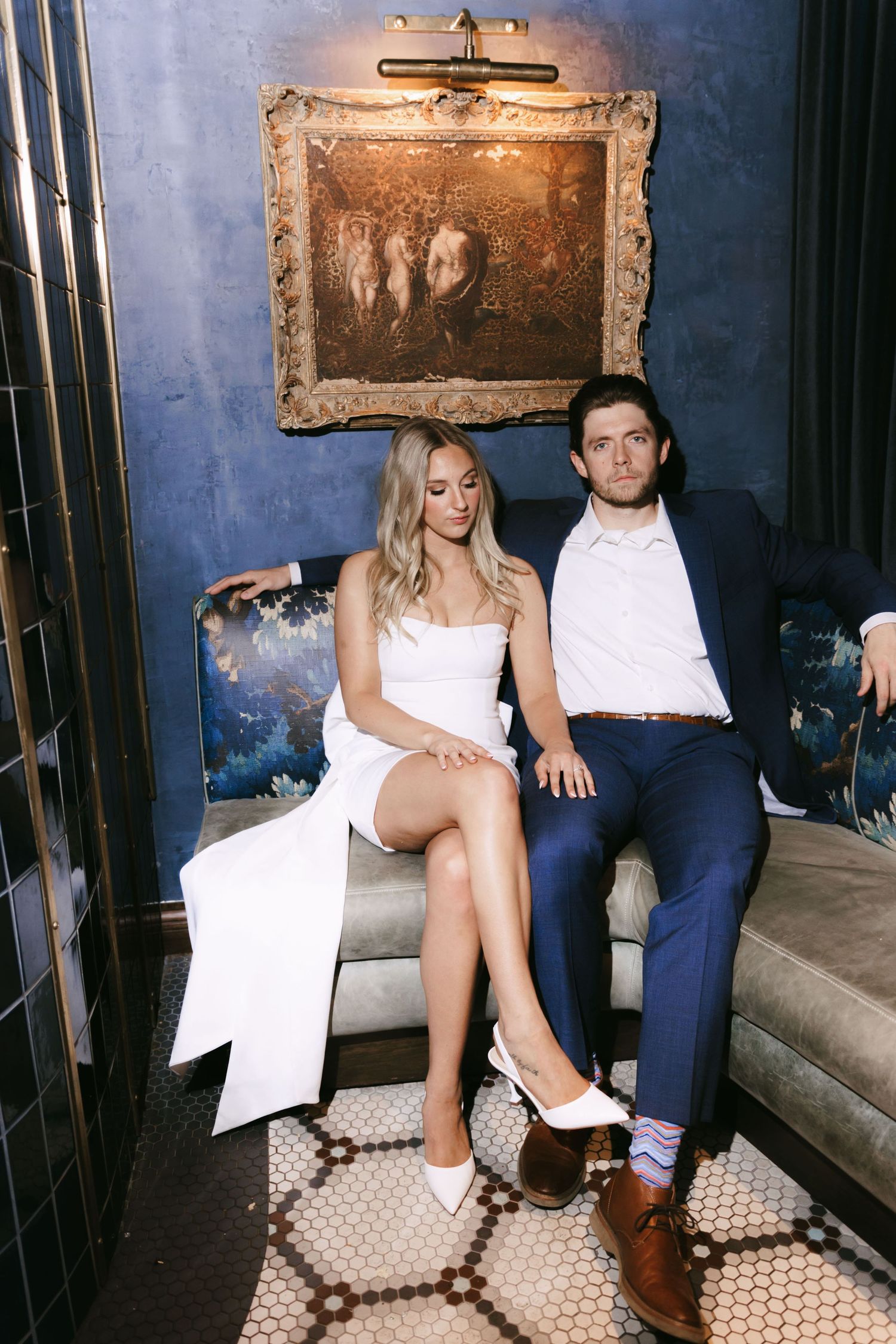 A stylish couple sits together on a vintage couch against a dark blue wall. Georgia state capitol wedding photographer.
